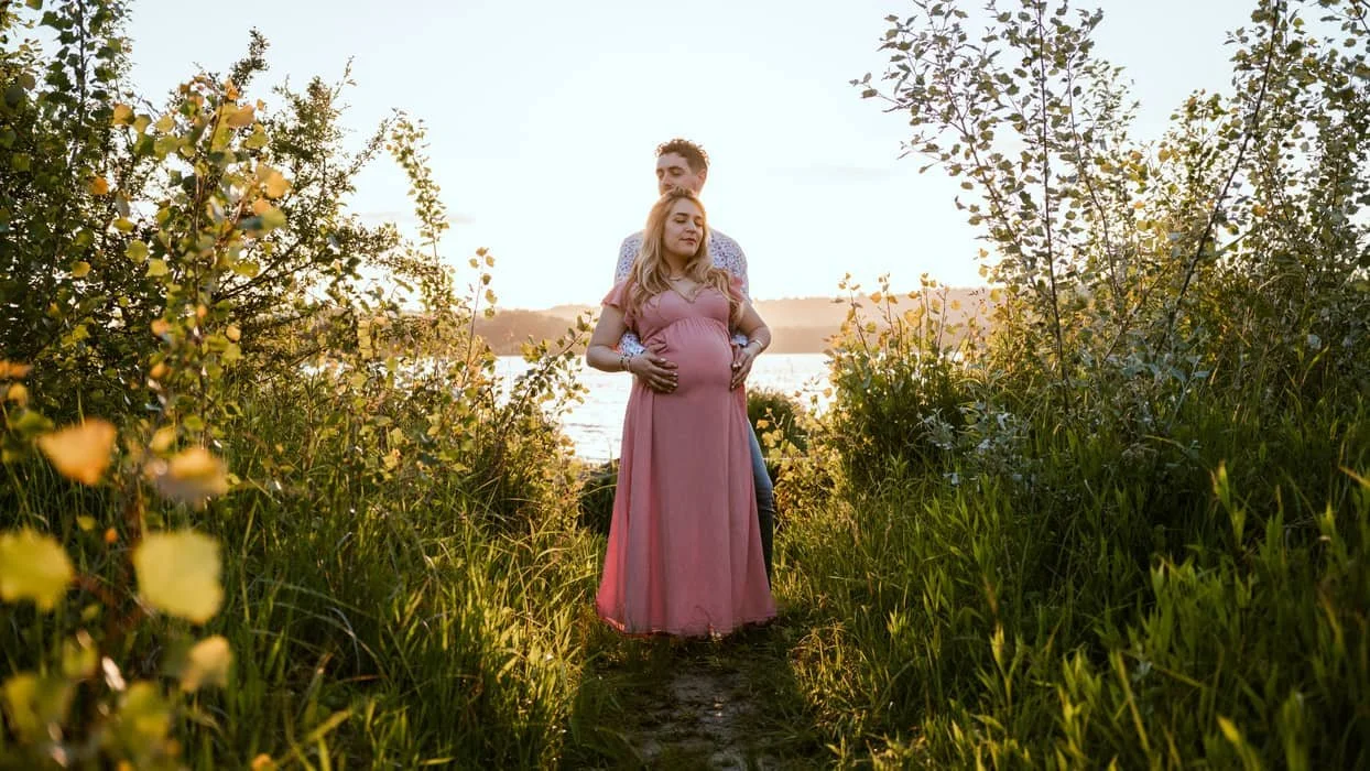 Une femme enceinte vêtue d'une robe rose, soutenue par un homme, dans un chemin naturel entouré d'arbustes et de végétation, avec un coucher de soleil en arrière-plan.