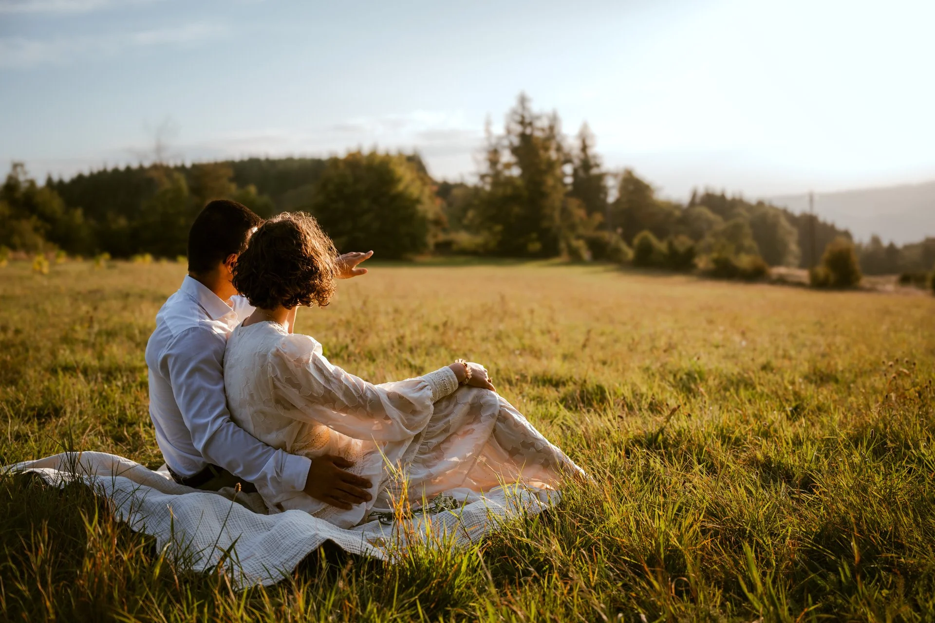 Un couple assis dans un champ d'herbes, regardant le coucher de soleil, entouré d'arbres et d'un ciel clair.