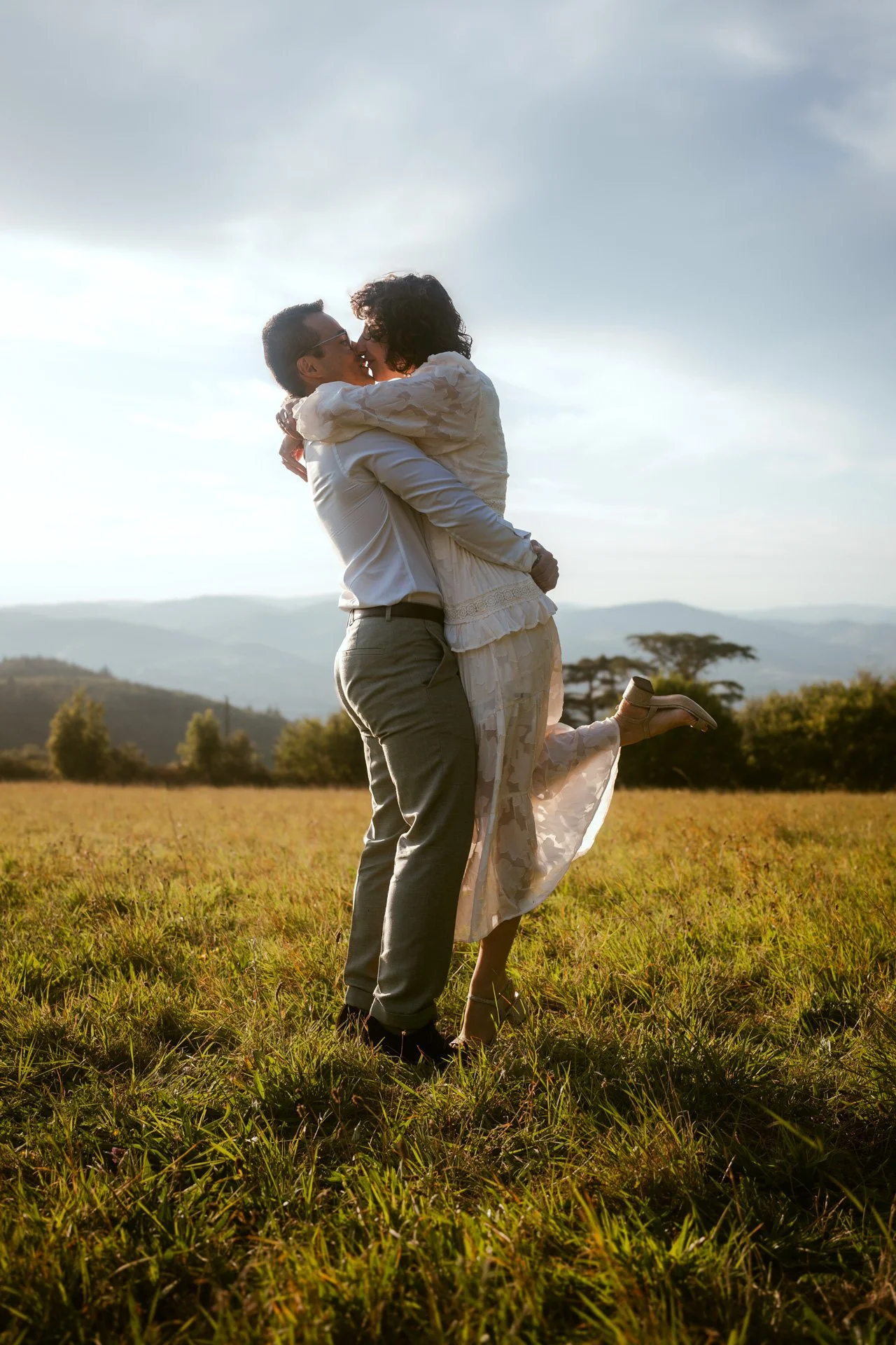 Un couple en vêtements élégants s'enlace dans un champ, avec un paysage de montagnes en arrière-plan, sous un ciel partiellement nuageux, lors d'une journée ensoleillée.