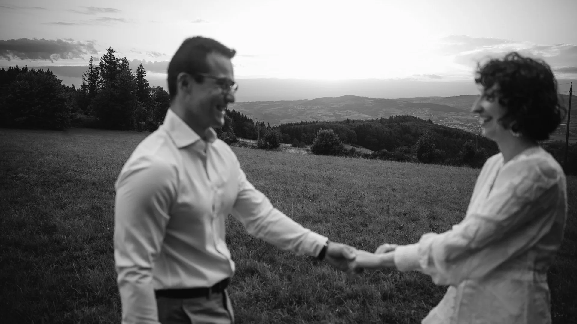 Un couple souriant se tenant la main dans un champ avec une vue de montagnes et de forêt en arrière-plan, lors du coucher du soleil ou du crépuscule.