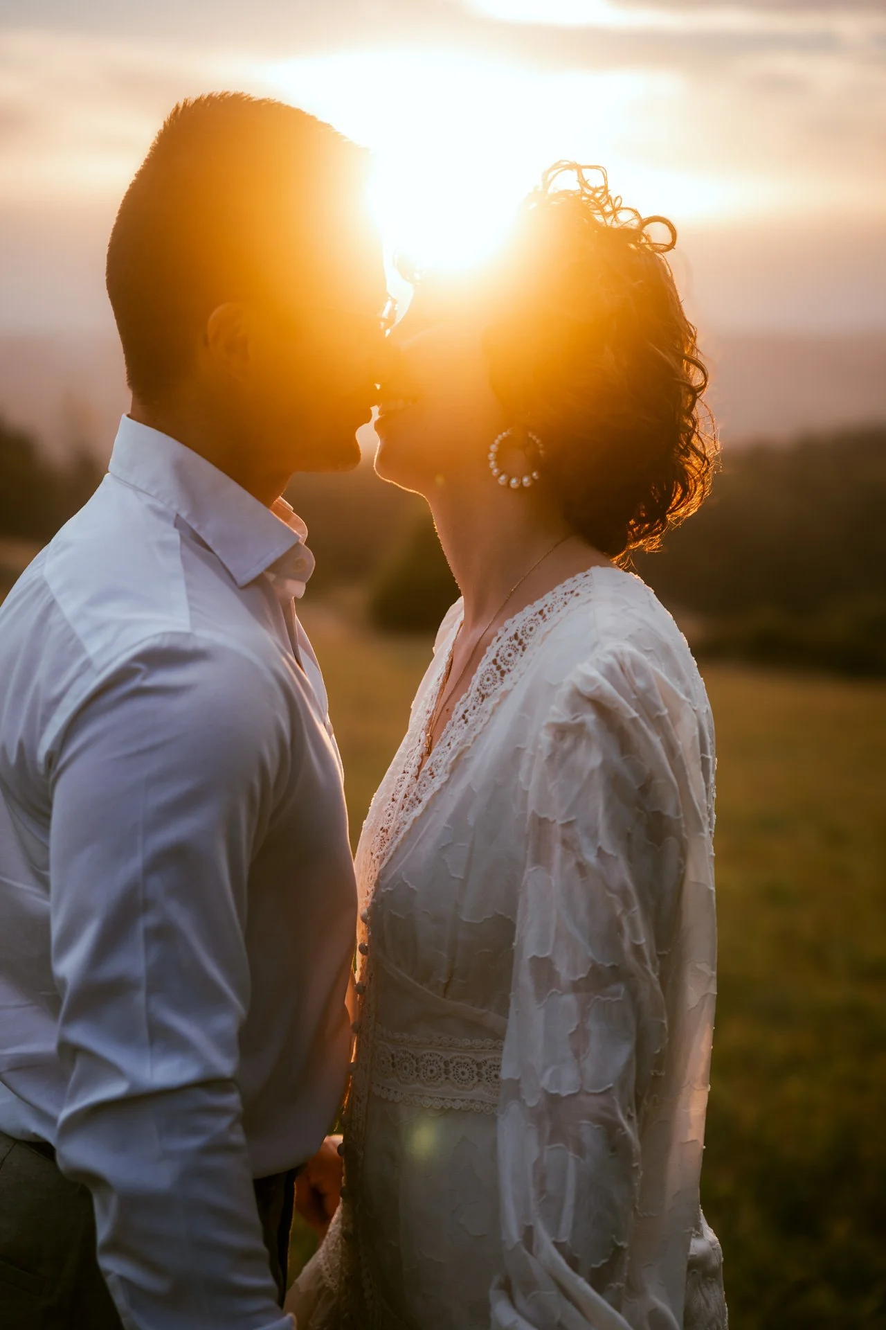 Un couple s'embrassant au coucher du soleil, en plein air, avec un ciel orange et un paysage flou en arrière-plan.