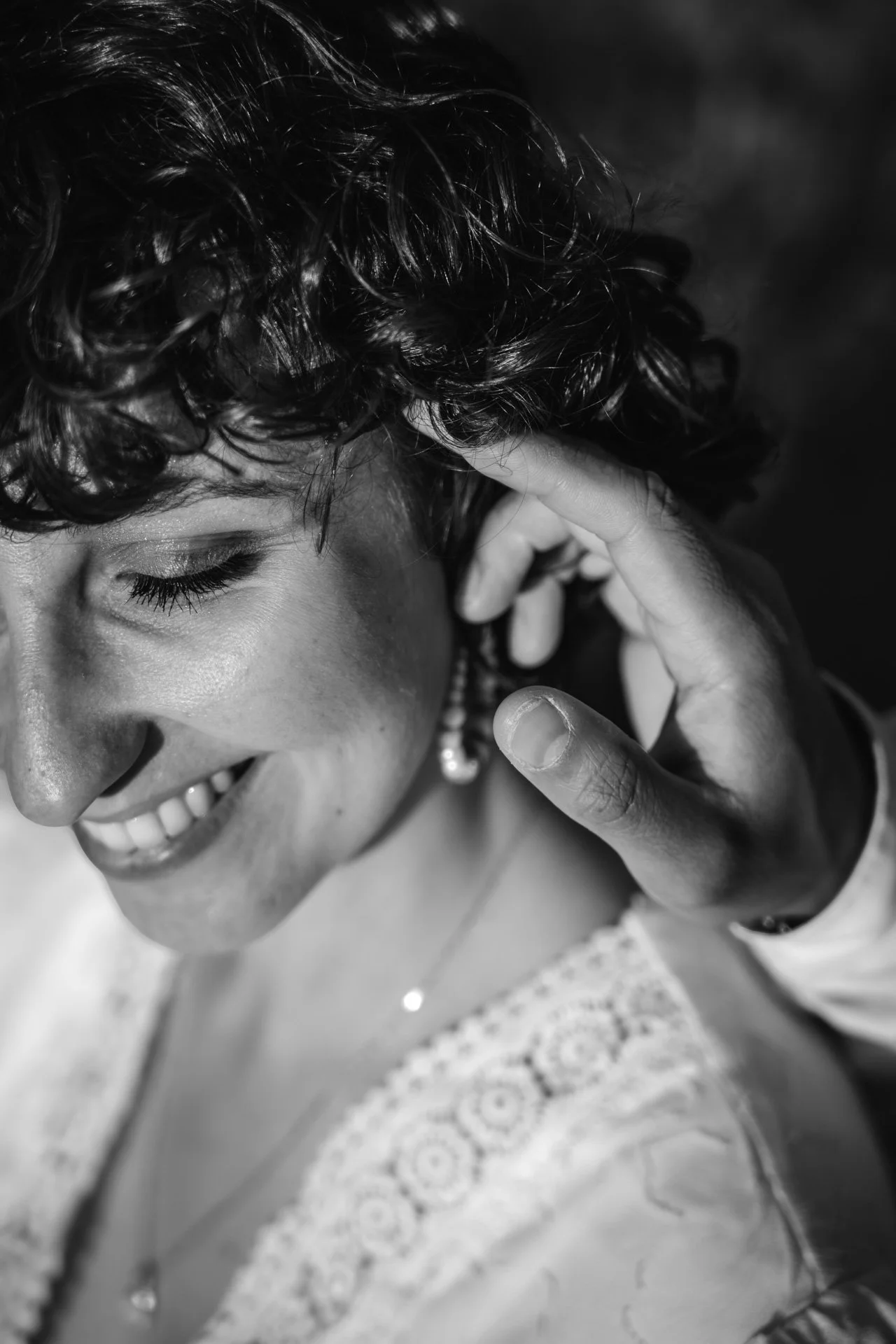Un portrait en noir et blanc d'une femme souriante avec des cheveux bouclés, portant une boucle d'oreille en perles et un collier, avec une main touchant son oreille.