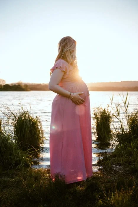 Femme enceinte en robe rose, debout au bord de l'eau lors du coucher de soleil, tenant son ventre