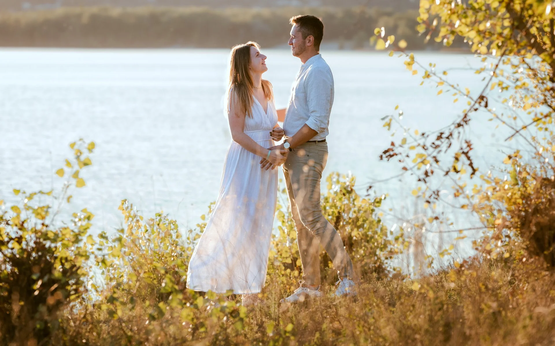 Un couple debout près d'un lac en été, se regardant tendrement, avec des arbres aux feuilles jaunes en arrière-plan.