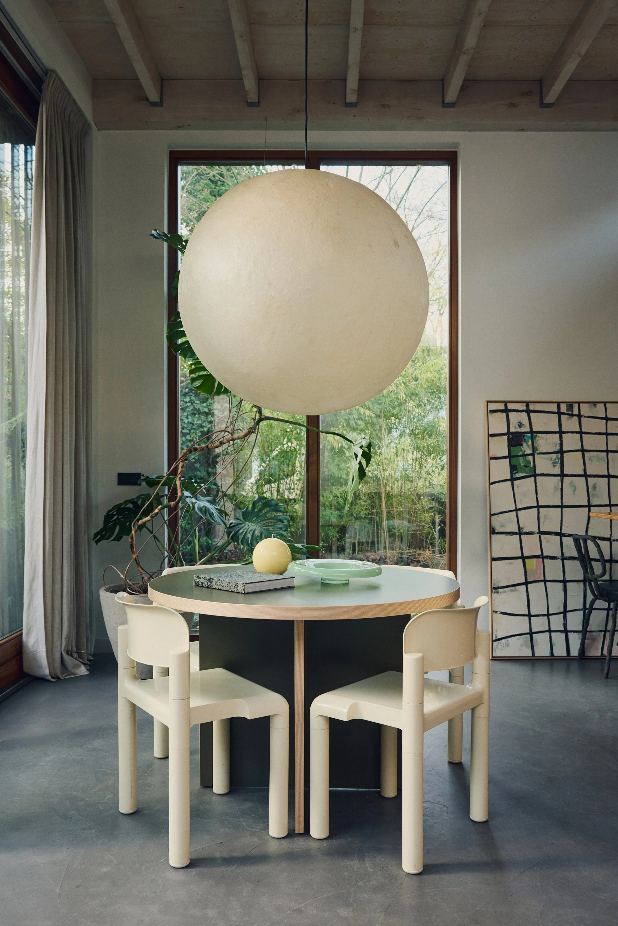 Eetkamertafel met vier moderne witte stoelen, decoratieve planten en een grote hanglamp in een lichte, natuurlijke binnenruimte met grote ramen en een kunstwerk aan de muur.