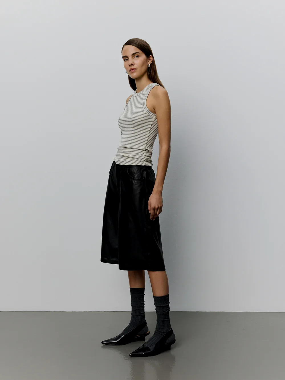 Een jonge vrouw met bruin haar, gekleed in een gestreepte tanktop en een zwarte, wijde rok, staat tegen een witmuur, met schoenen en sokken, in een minimalistische setting.