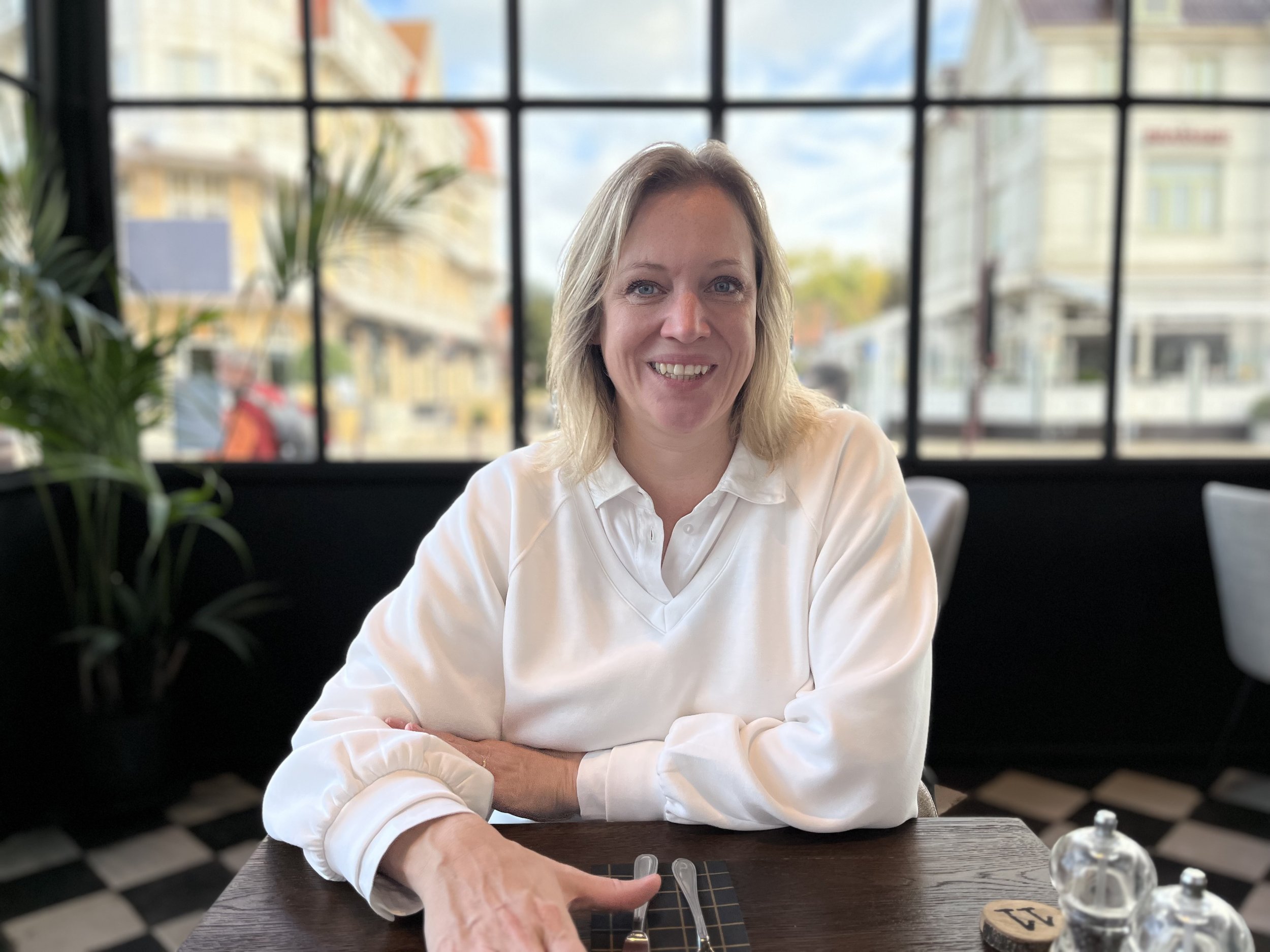 Vrouw zit aan een tafel in een restaurant, lachend naar de camera, met grote ramen en een stedelijke straat op de achtergrond.