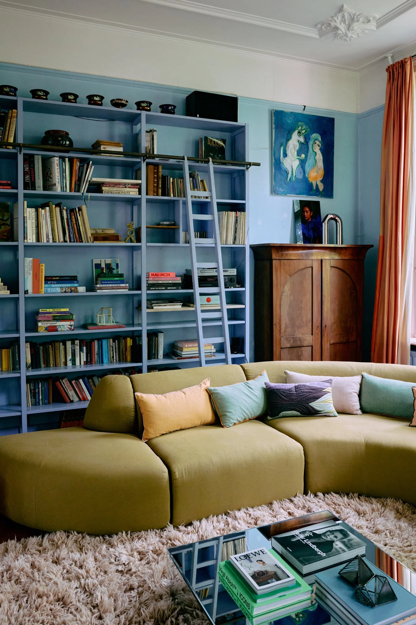 Gezellig woonkamer met een hoekbank in olijfkleur, omgeven door kleurrijke kussens, zachte vloerkleed en een salontafel met boeken en decoraties. Achter de bank een grote blauwe boekenkast vol boeken en decoratieve voorwerpen, met een ladder om boeken te bereiken. Aan de rechterkant een houten kast en een raam met oranje gordijnen. Boven de kast hangen twee kunstwerken, een kleurrijk schilderij en een foto.