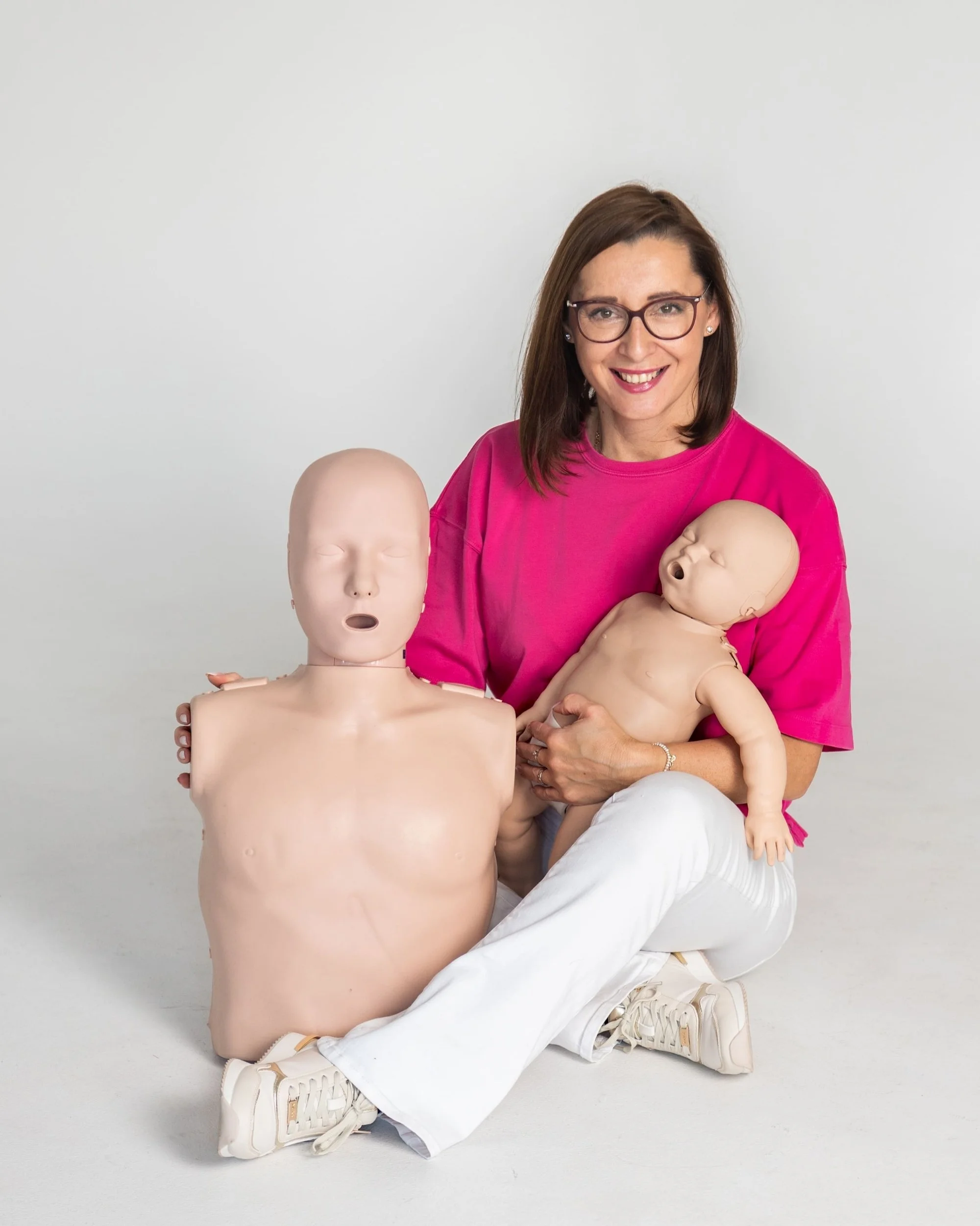 A woman with brown hair, glasses, and a bright pink shirt kneeling on the floor, holding a beige baby doll in her arms, with two other dolls nearby.