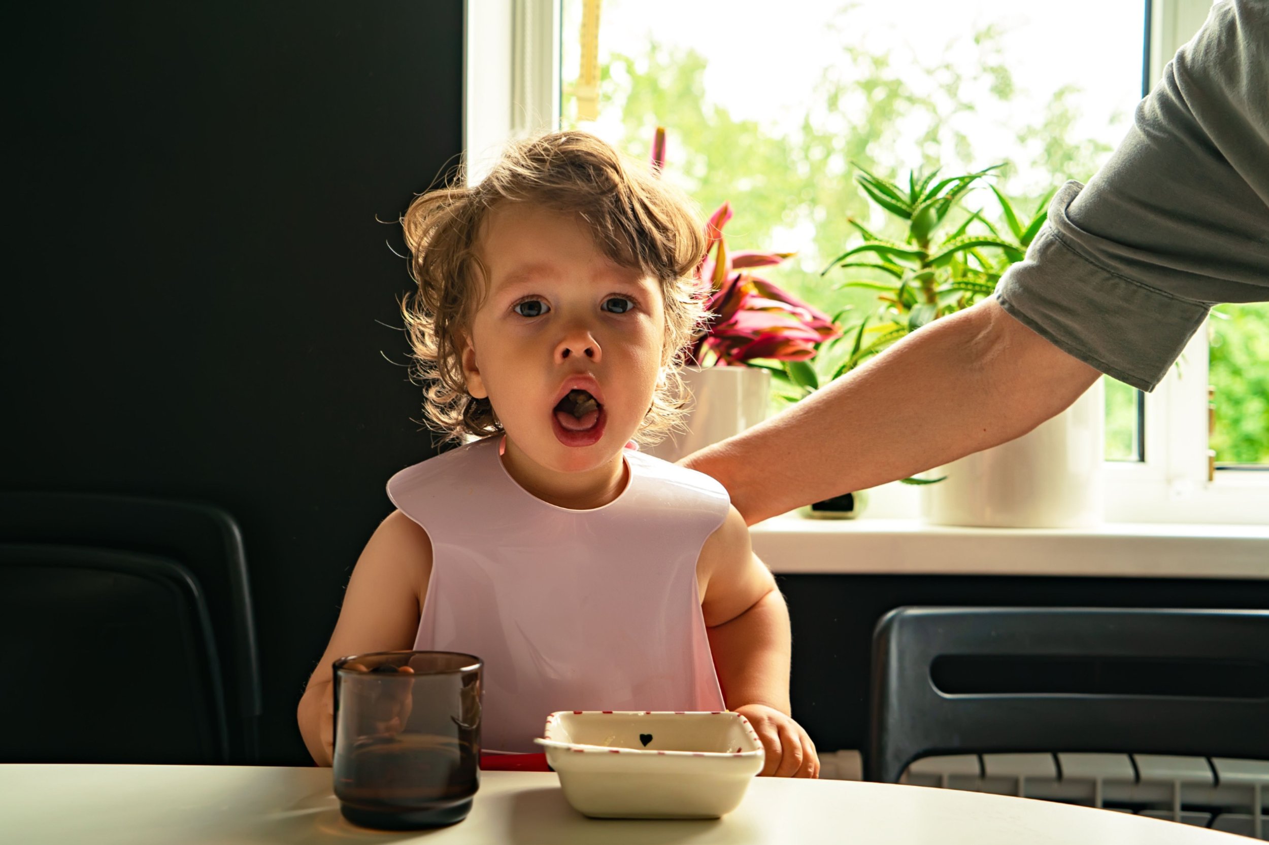 Young girl with curly hair and a pink sleeveless top sitting at a table with a glass of water, a bowl, and a tray in front of her, with a window and plants in the background.