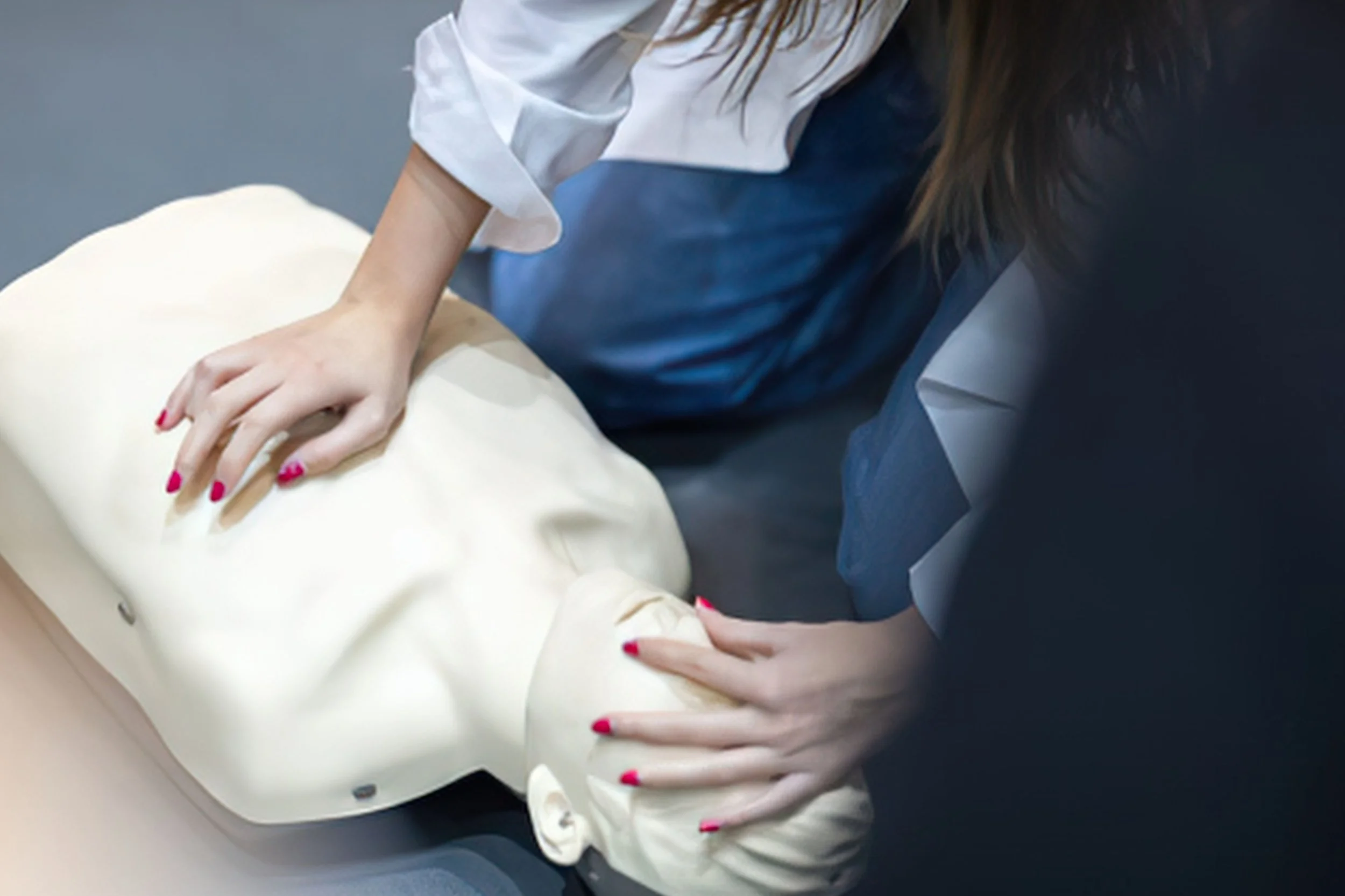 A person with long brown hair and painted red nails is performing CPR on a mannequin, pressing down on its chest with both hands. The mannequin is dressed in a cream-colored top, and the person is wearing a navy blue uniform or outfit.