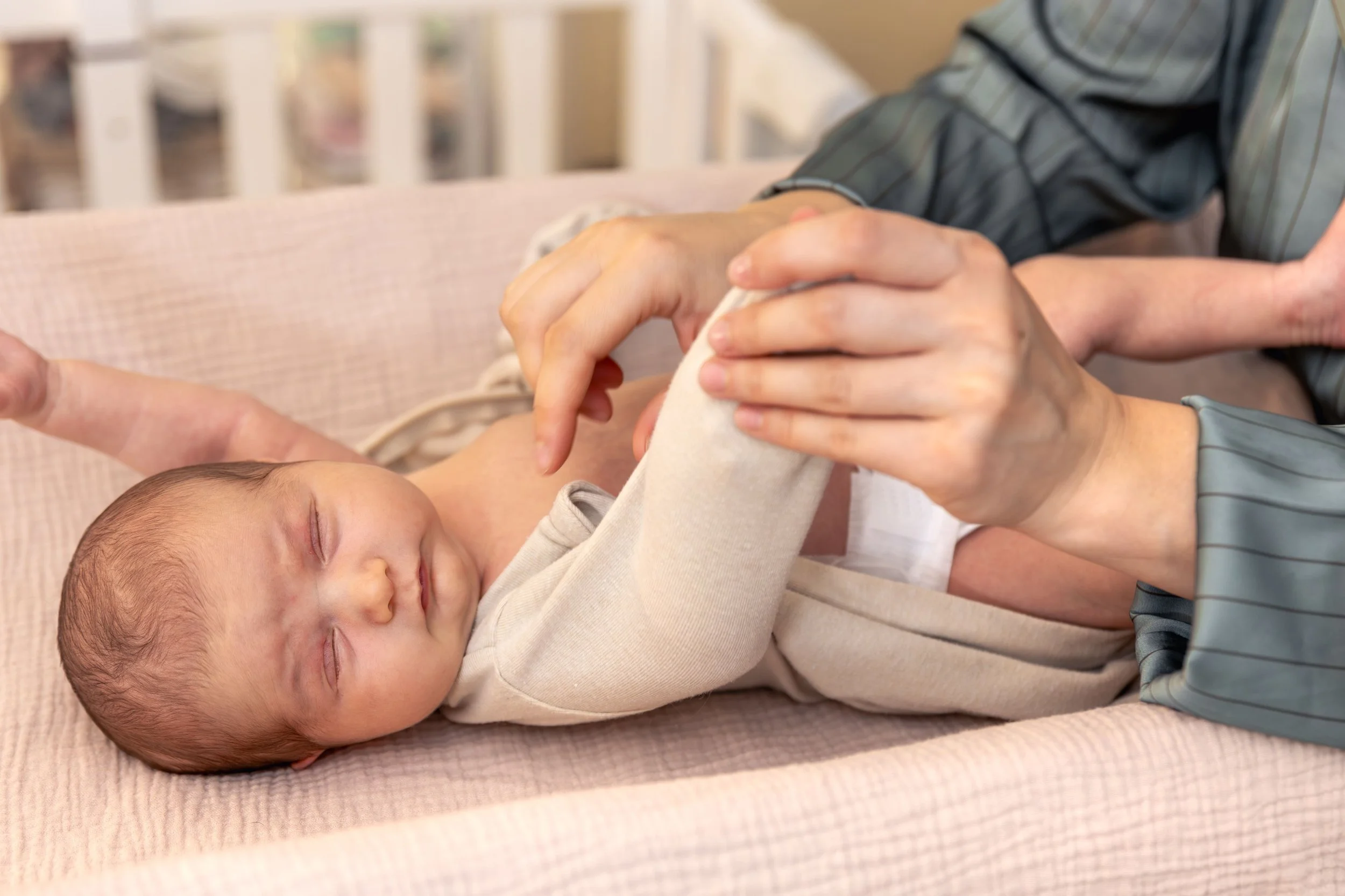 A baby lying on a pink blanket with closed eyes, holding hands with an adult whose face is not visible.