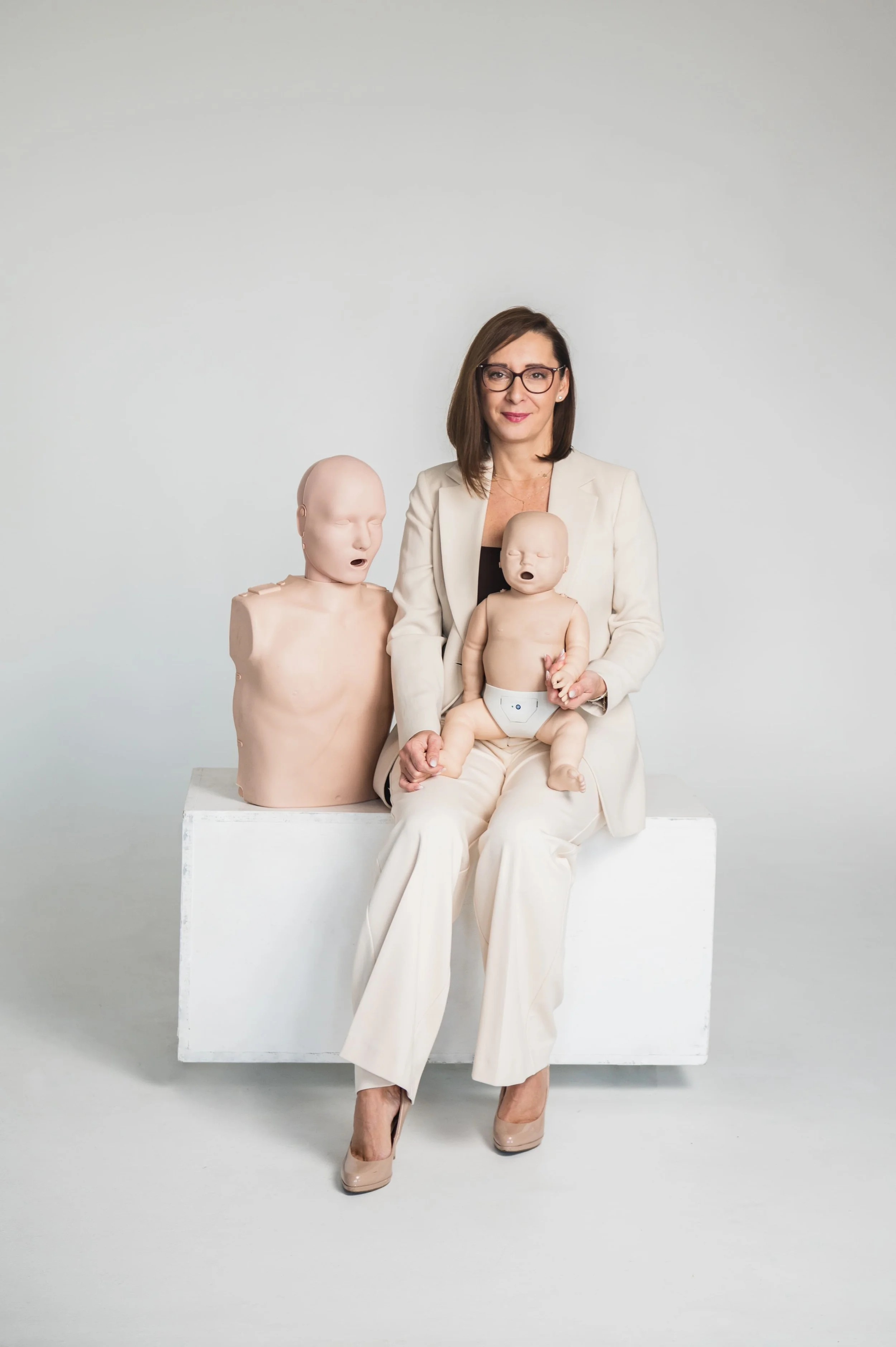 A woman in a cream-colored suit and heels sitting on a white block, holding a crying baby doll, with two other crying baby dolls standing on either side of her, against a plain gray background.