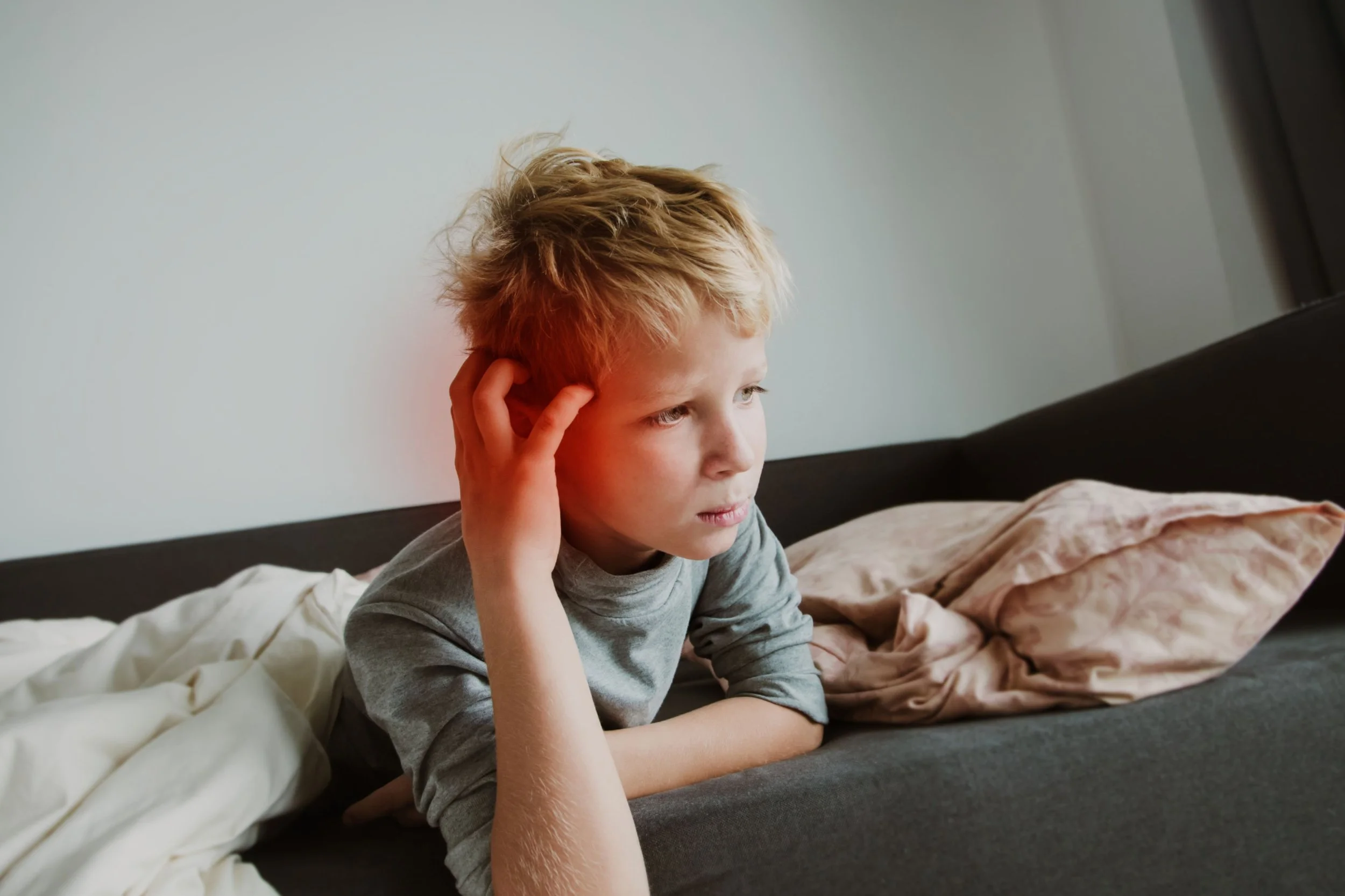 A young boy with blond hair is sitting on a bed, looking at a screen with a serious expression. He is touching his head with his left hand, and the bed has rumpled sheets and a pillow.