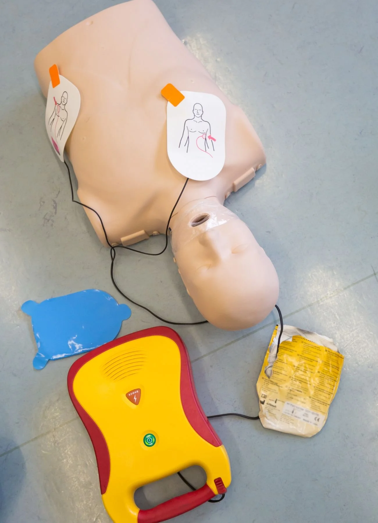 A medical training mannequin with connected wires for CPR practice, with instructional stickers, a blue pad, and a yellow AED trainer device on the floor.