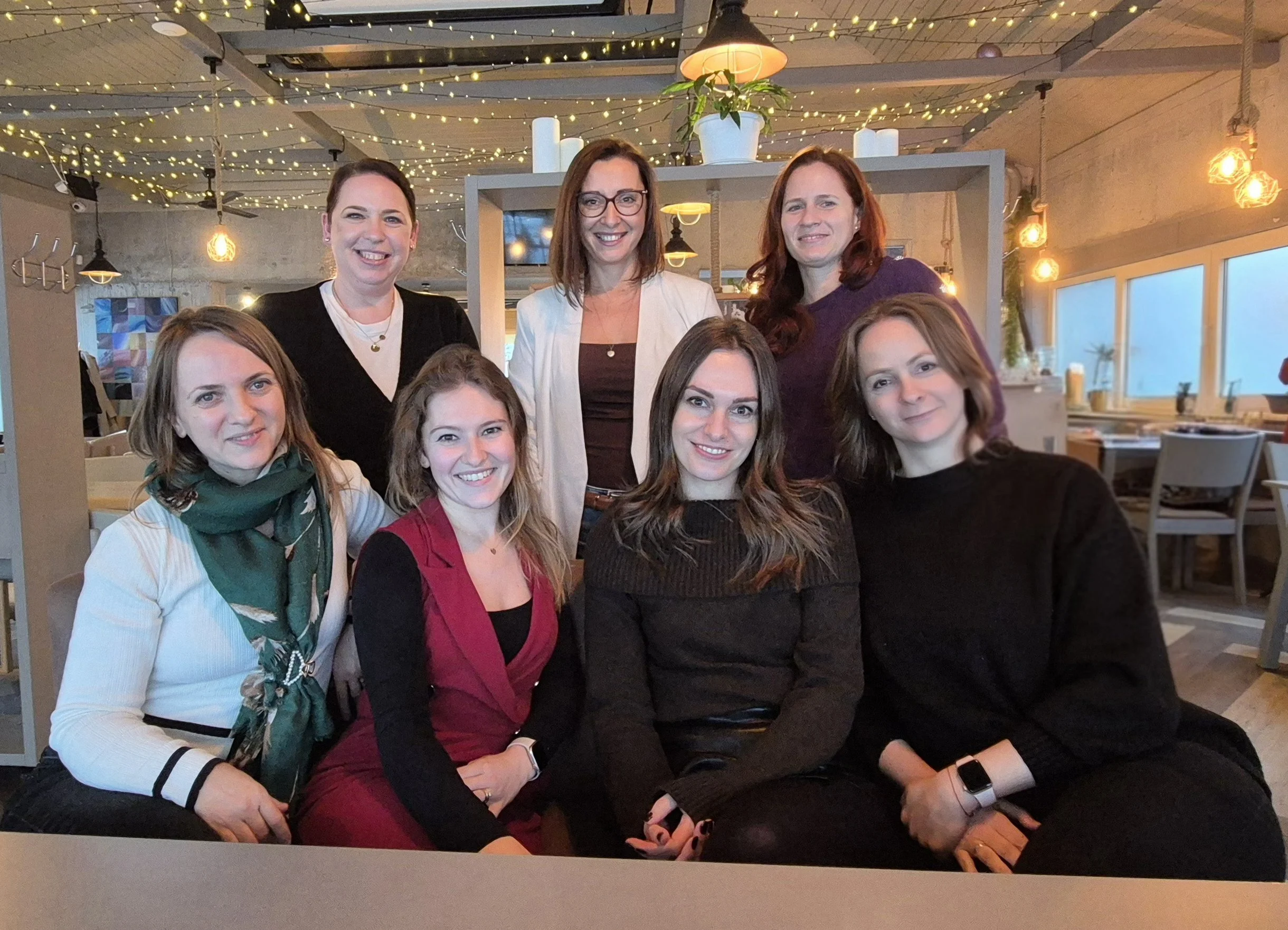 A group of nine women sitting and standing together inside a warmly lit restaurant or cafe, smiling at the camera, with string lights and modern decor overhead.