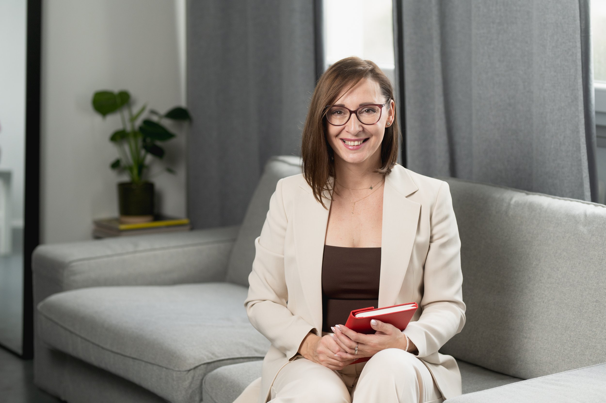 A smiling woman with brown hair, glasses, and a beige blazer sitting on a light gray sofa, holding a red notepad in her hands. There is a potted plant and some magazines in the background.