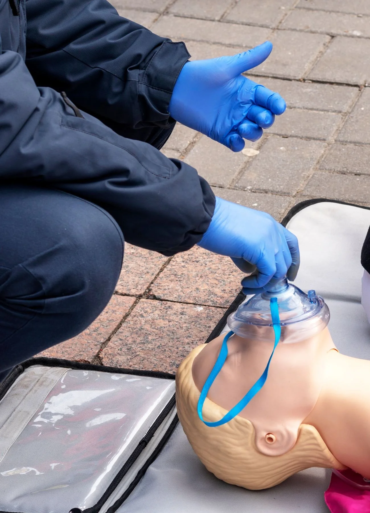 A person in blue gloves and dark clothing practicing CPR on a training mannequin outdoors on a brick surface.