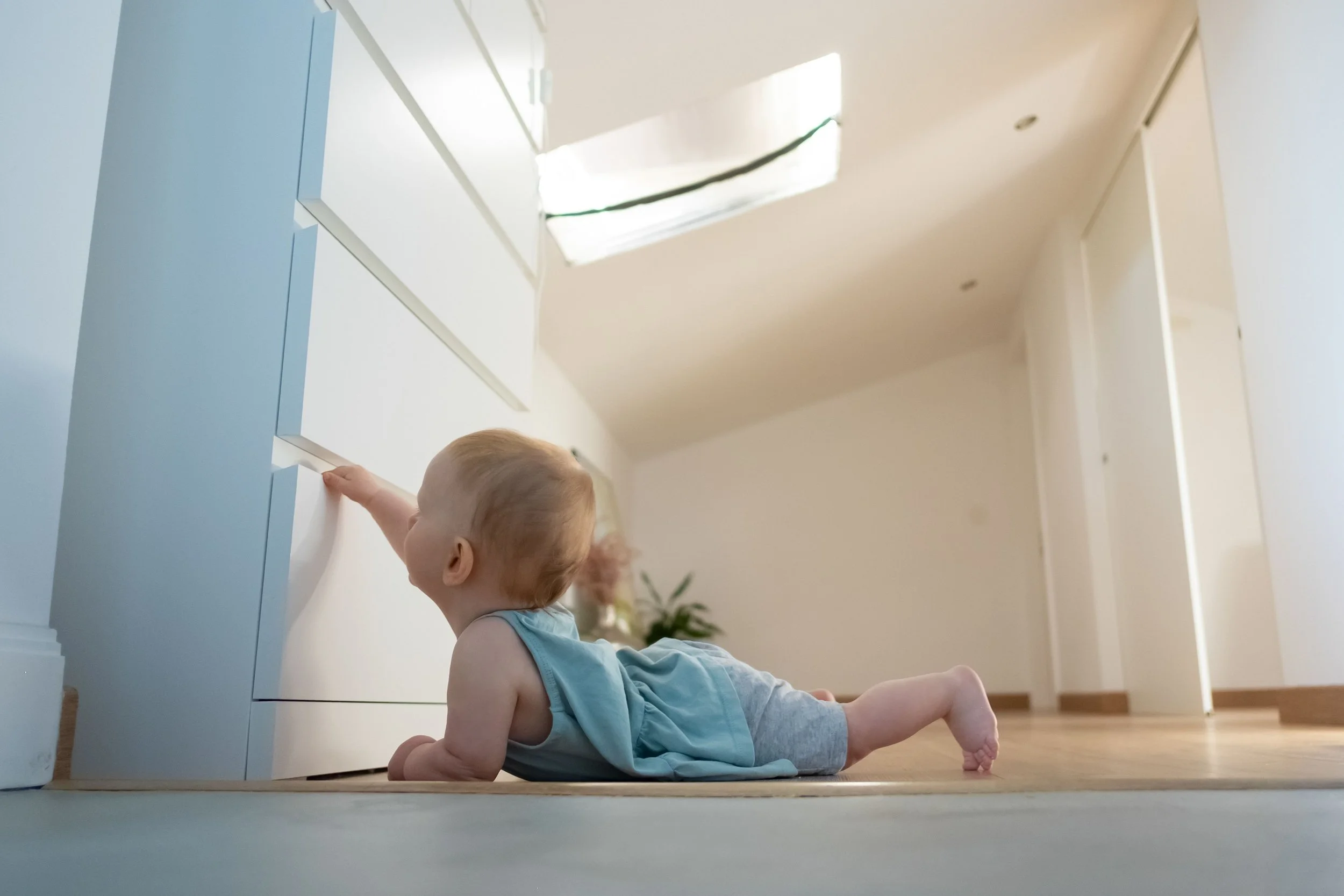 A young child with light hair, wearing a light blue sleeveless dress, is lying on their stomach on a wooden floor, reaching up to a white kitchen cabinet.