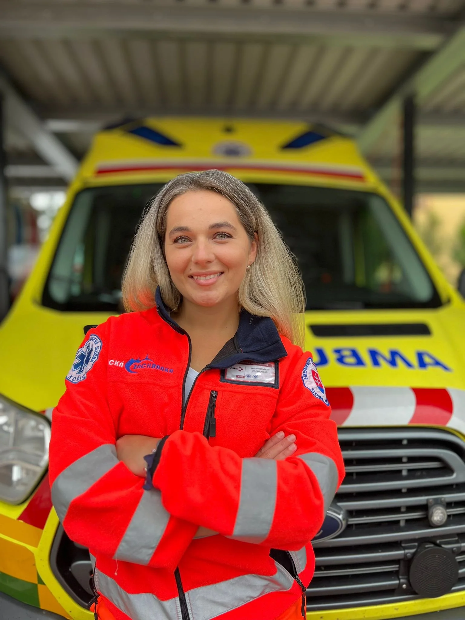 A smiling woman in a red emergency response jacket standing with arms crossed in front of an ambulance.