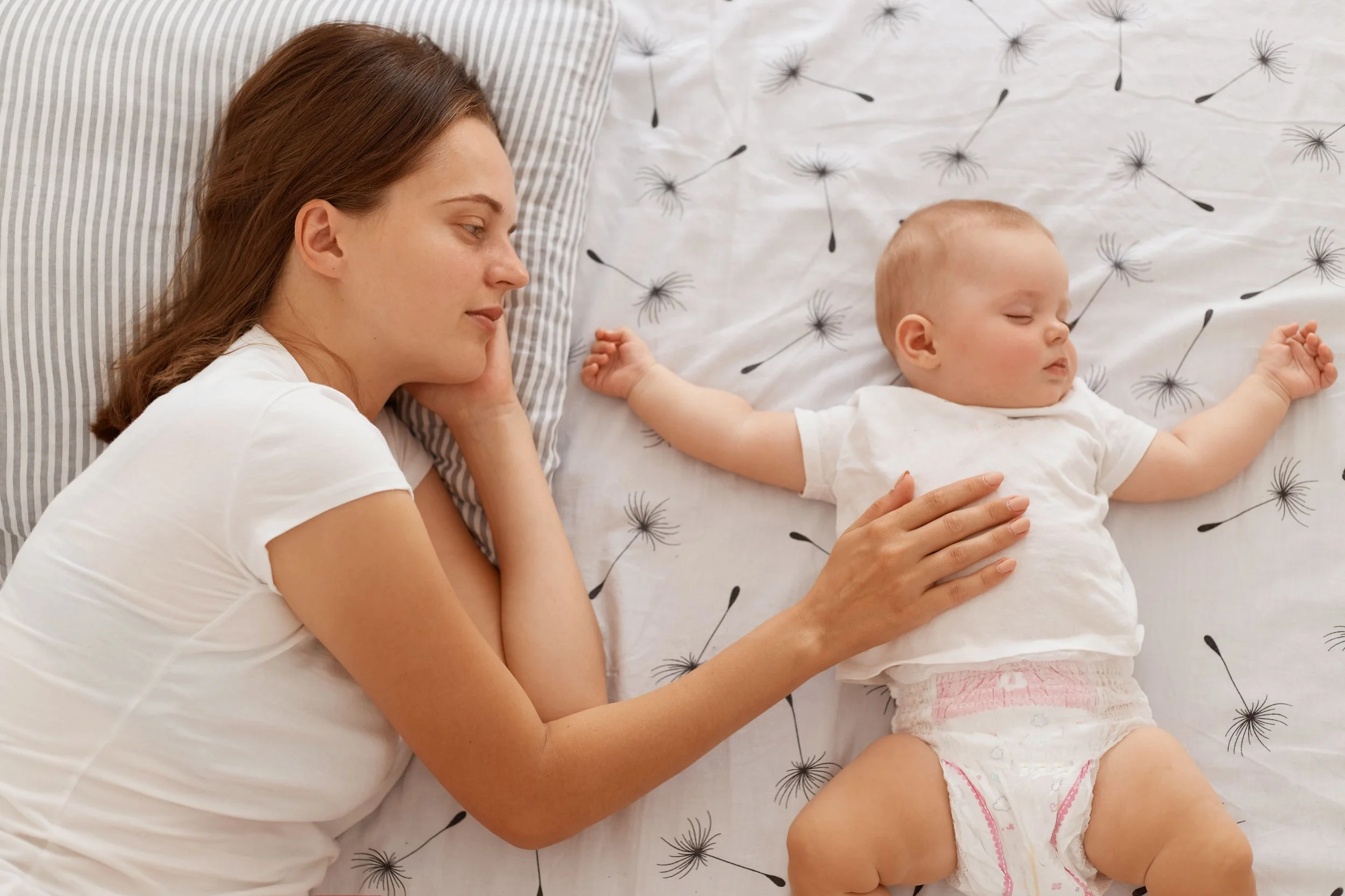A woman and a baby lying on a bed, both with eyes closed. The woman is resting her head on a striped pillow and gently holding the baby with her hand. The bed has a white sheet with dandelion patterns.