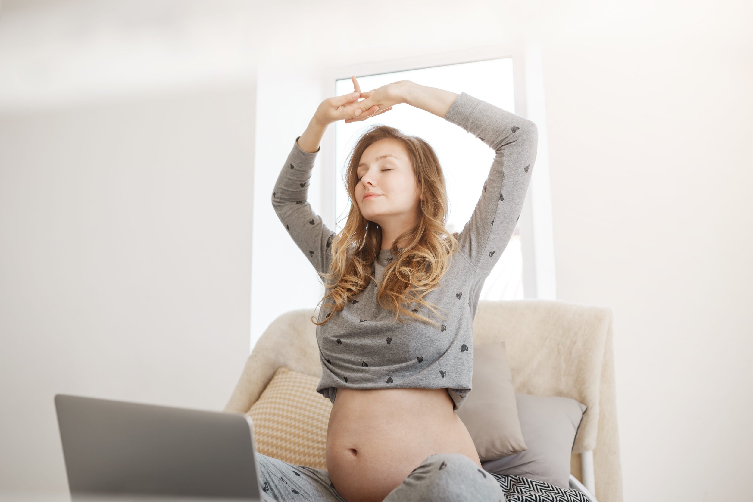 A young woman with long, wavy blonde hair and closed eyes, wearing a gray long-sleeve shirt with black hearts, is sitting on a couch with her eyes closed and arms raised above her head, in a bright, minimalistic room with a window behind her.