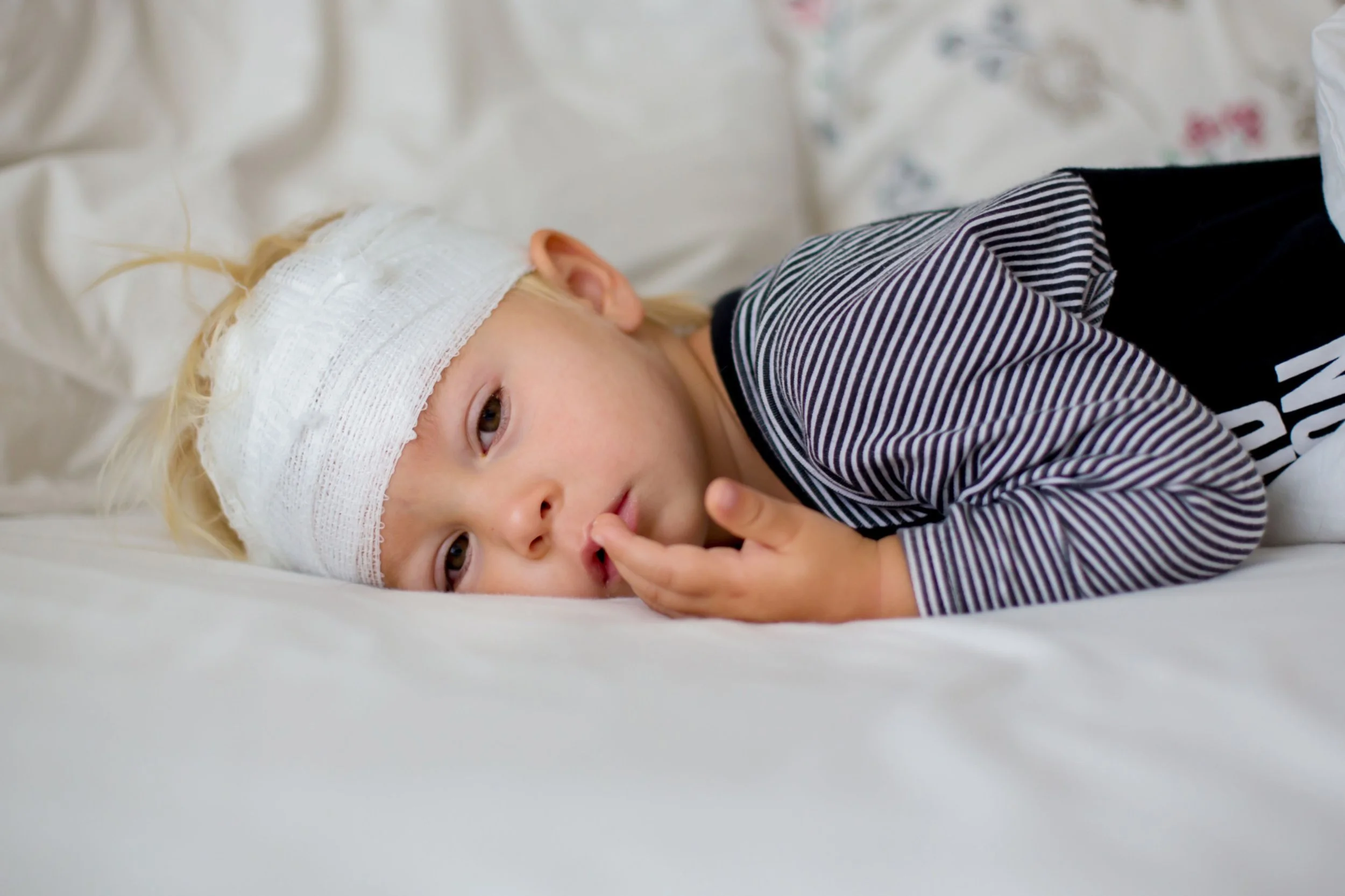 A young child lying in a hospital bed with a head injury, wearing a white bandage on their forehead, and resting on a white pillow.