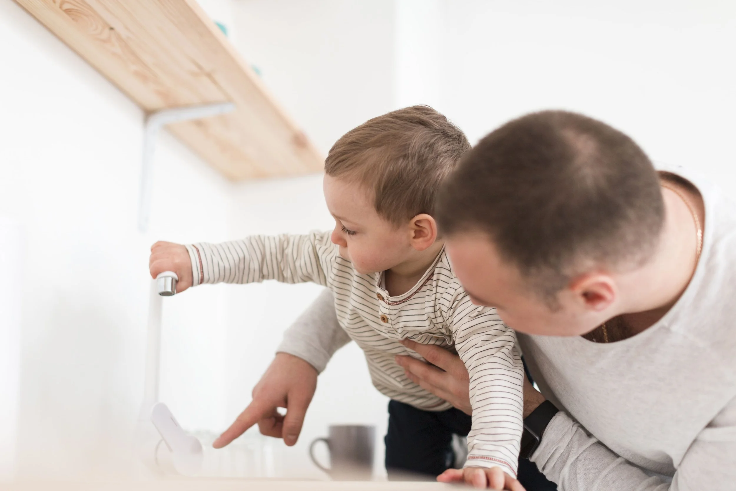 A young boy leaning over a white countertop with an adult, pointing at something on the surface, in a bright, minimal kitchen or room.
