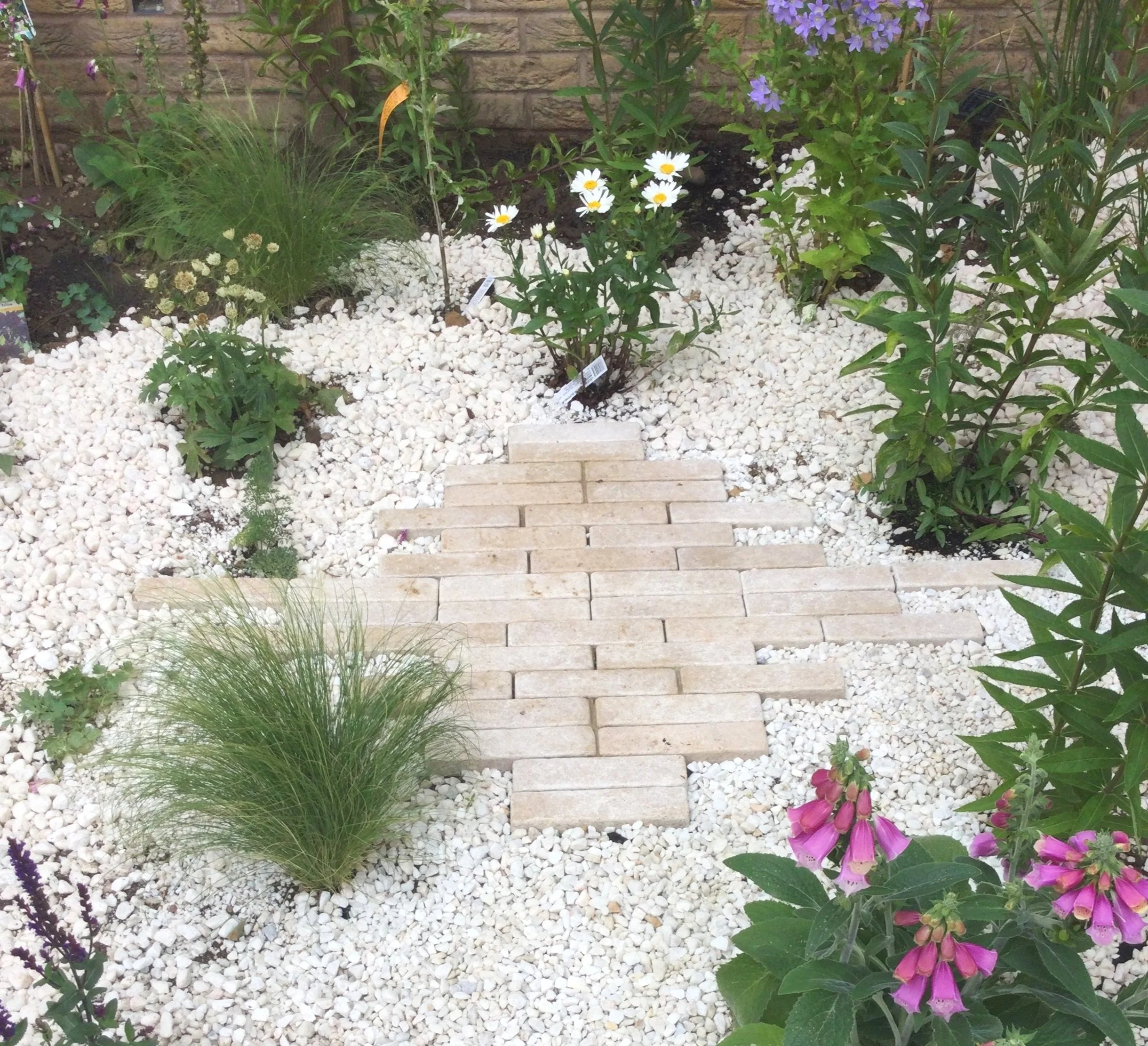 Small brick pathway leading through a garden with various flowering plants, including daisies, pink, purple, and white flowers, surrounded by white gravel.
