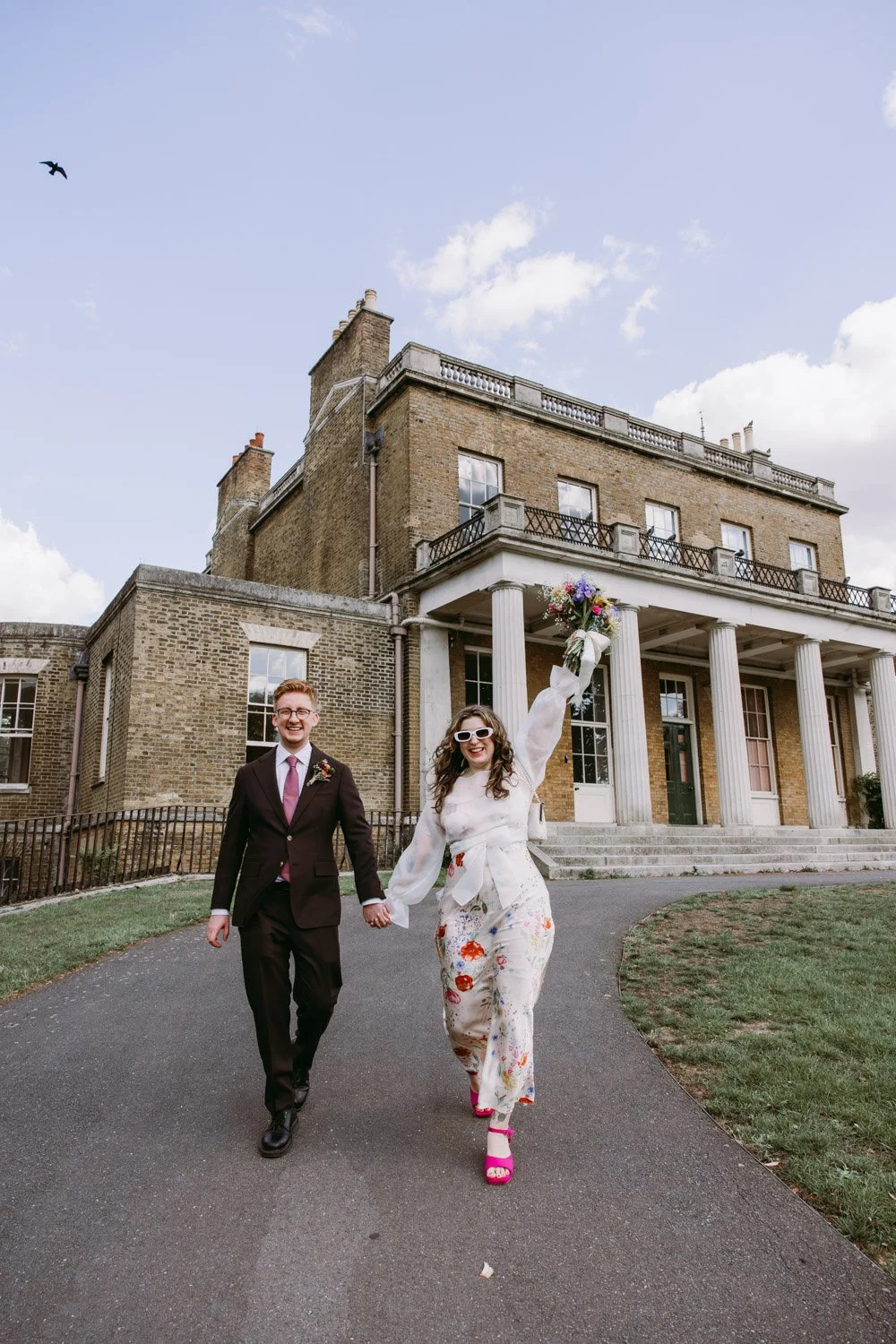 A joyful couple holding hands and walking outside a historic building, with the woman holding a bouquet and wearing colorful heels, celebrating a wedding or special event.