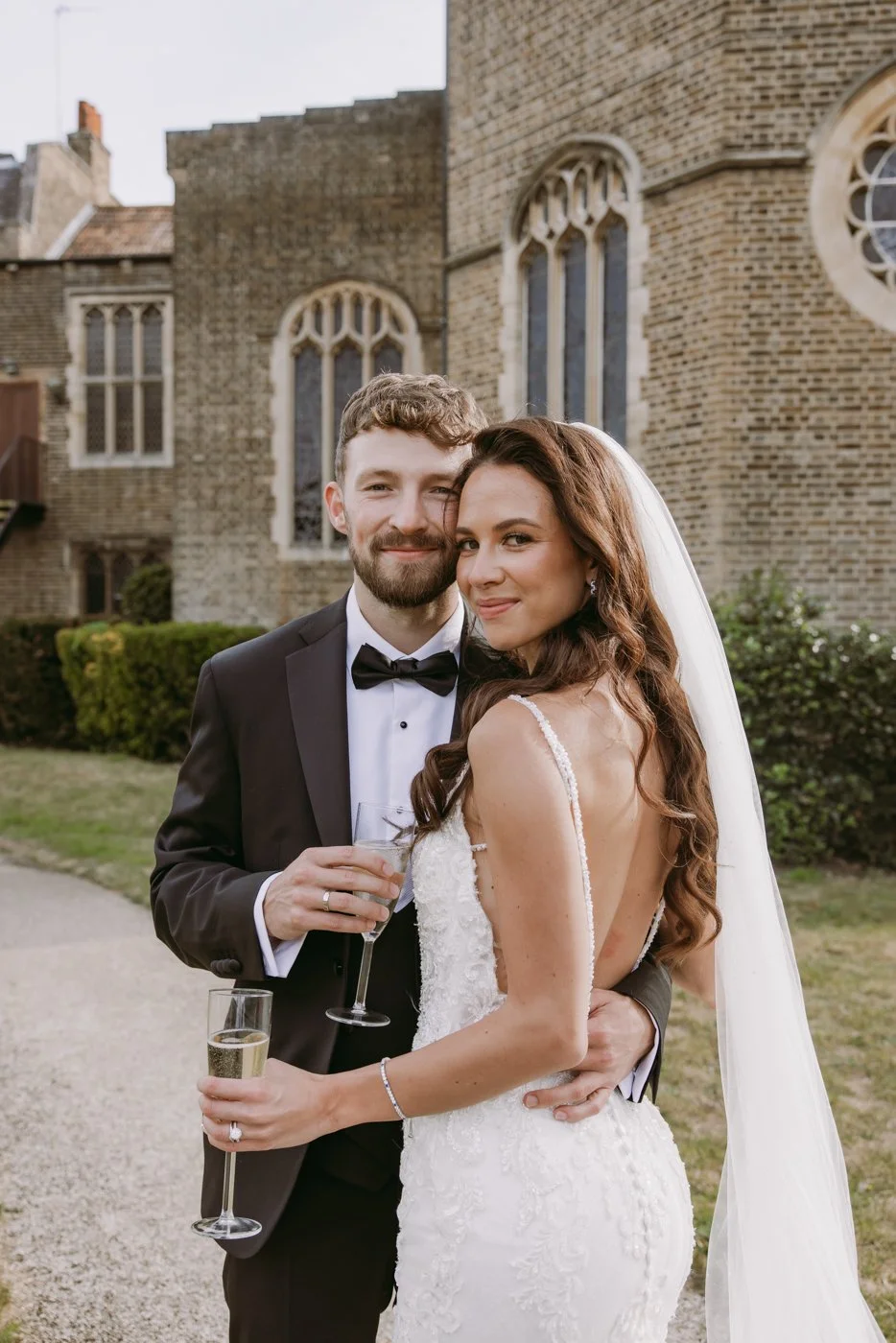 A bride and groom in wedding attire standing outside near a brick church, holding champagne glasses, smiling at the camera.