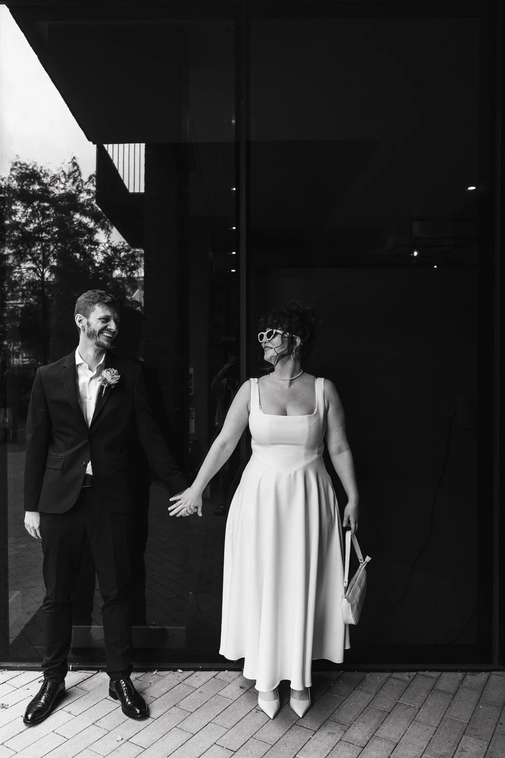 A black and white photo of a couple dressed in formal wedding attire, holding hands and smiling at each other outside a building with a glass facade.