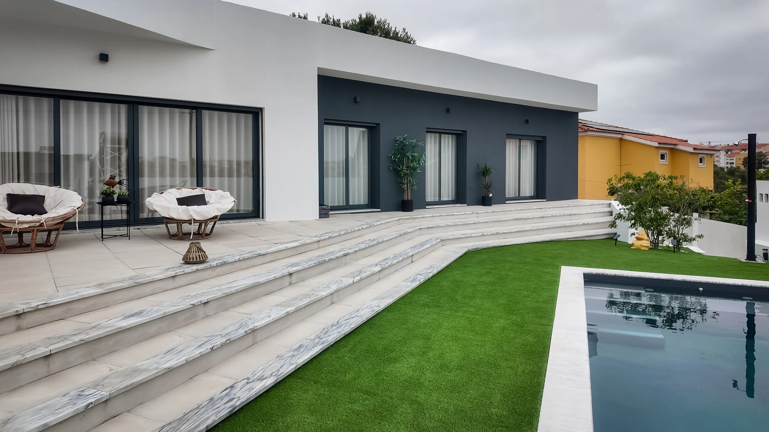 Casa moderna com jardim, escadas, área de churrasqueira com espreguiçadeiras e piscina, jardim bem cuidado, céu nublado.