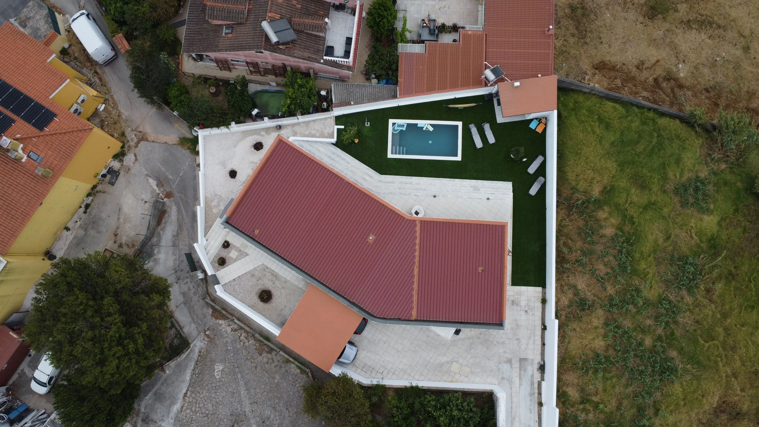 Imagem aérea de uma casa com telhado vermelho, piscina pequena, área com gramado e espaço de lazer ao ar livre, cercada por muro branco, em bairro residencial.