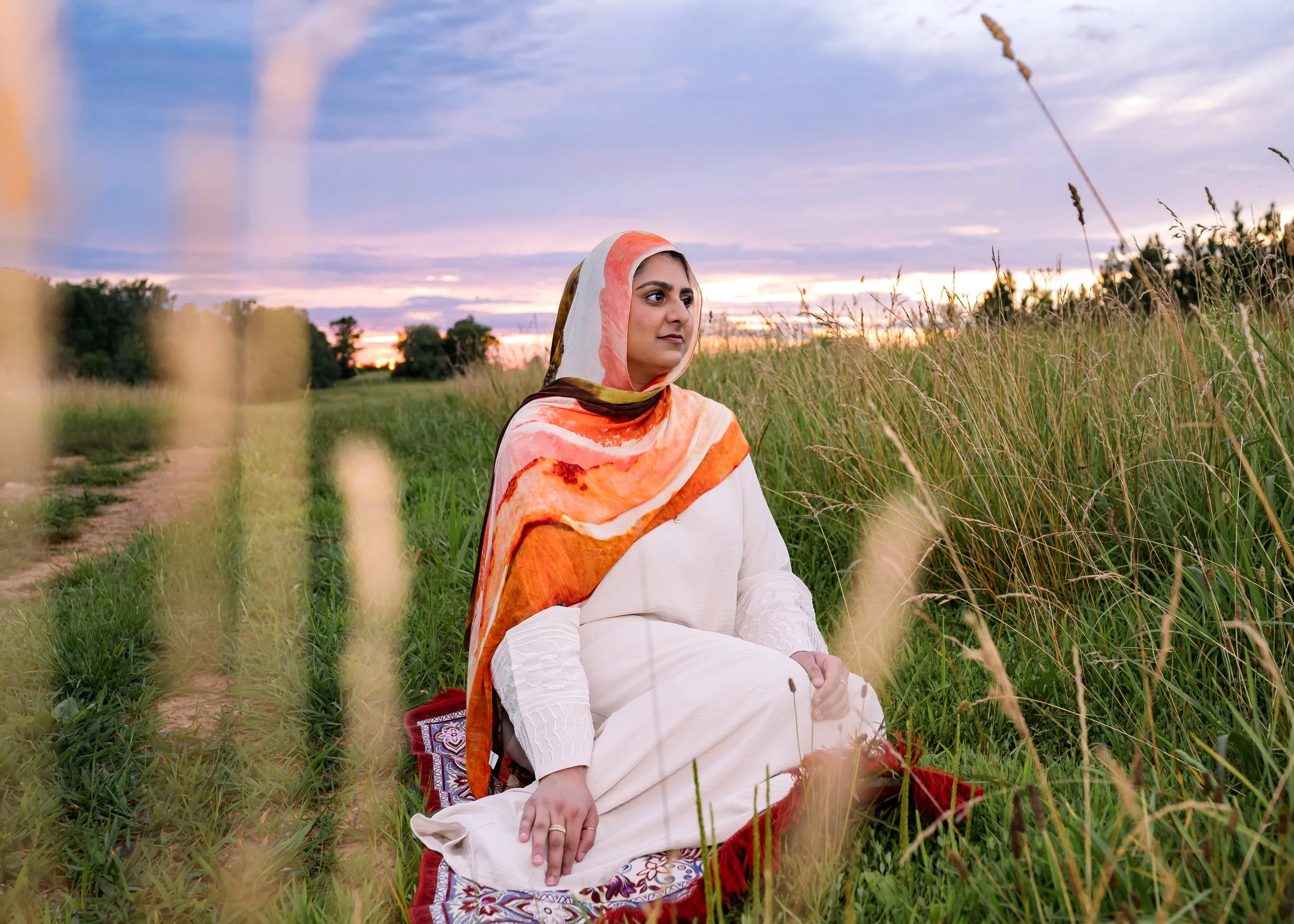 Hijabi sitting on a prayer mat in a field with a sunset in the background