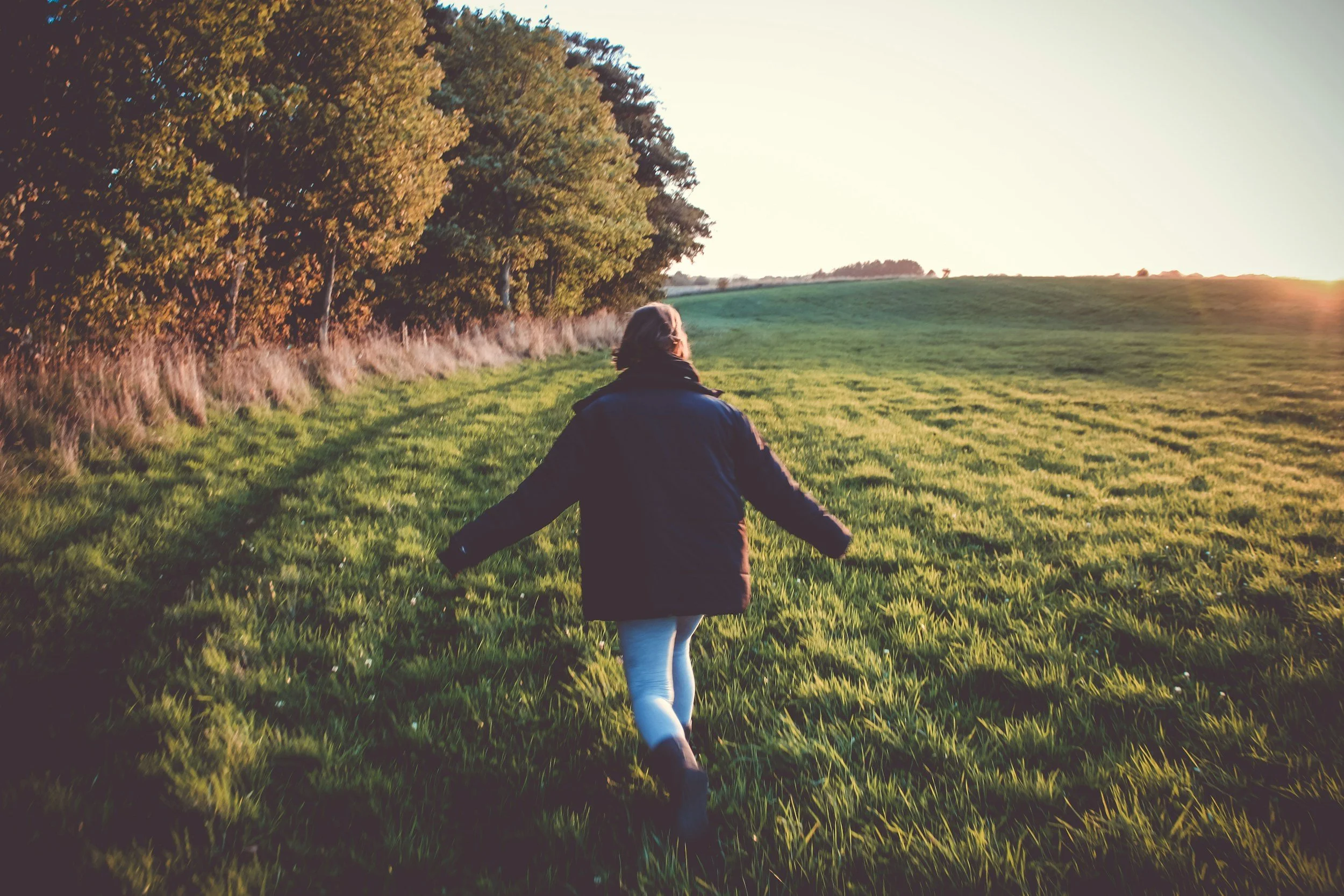 A woman walking through a grassy and sunny field with her arms out