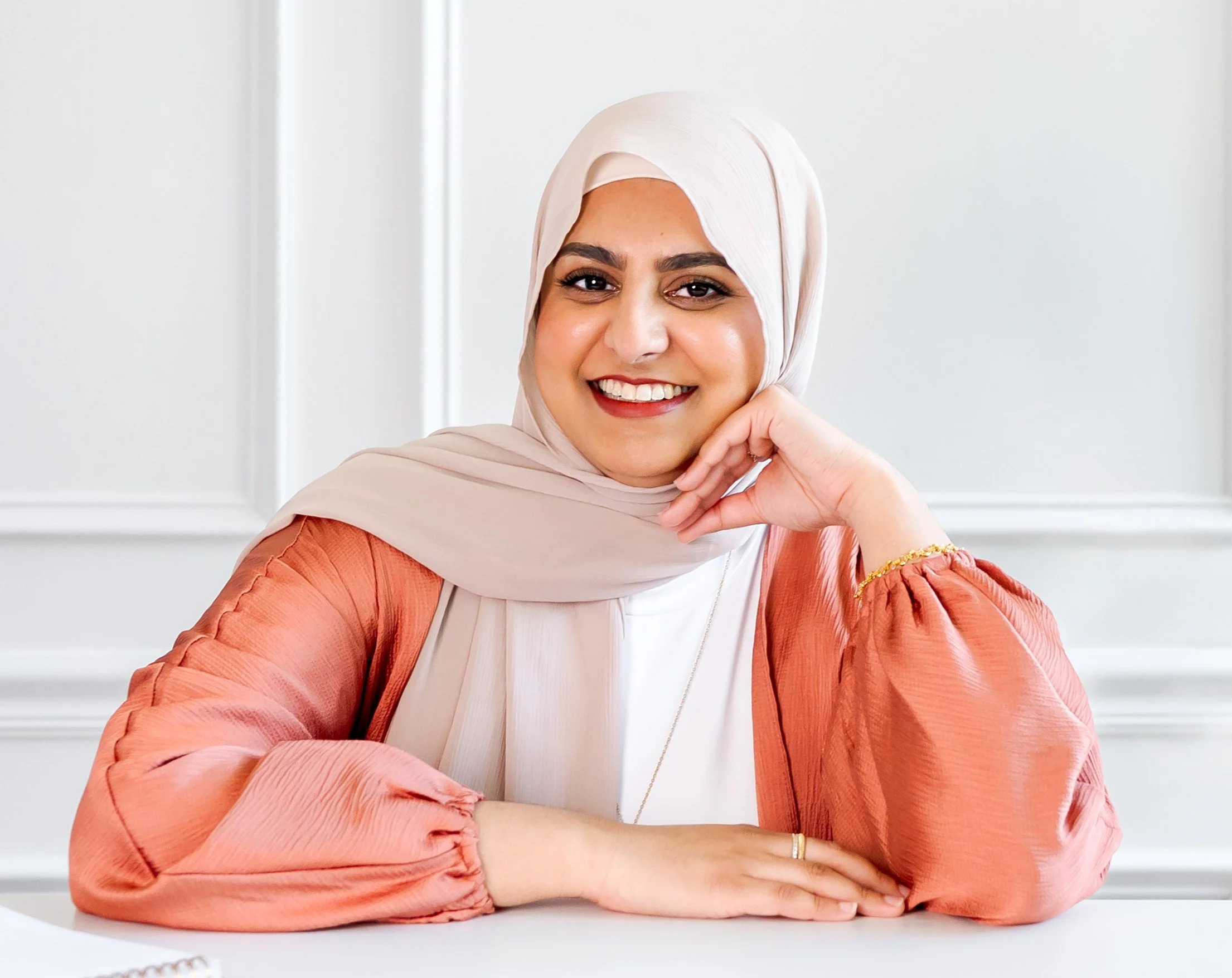 A woman wearing a light beige hijab and an orange top with puffy sleeves, smiling and resting her chin on her hand, in a white room with decorative wall molding.