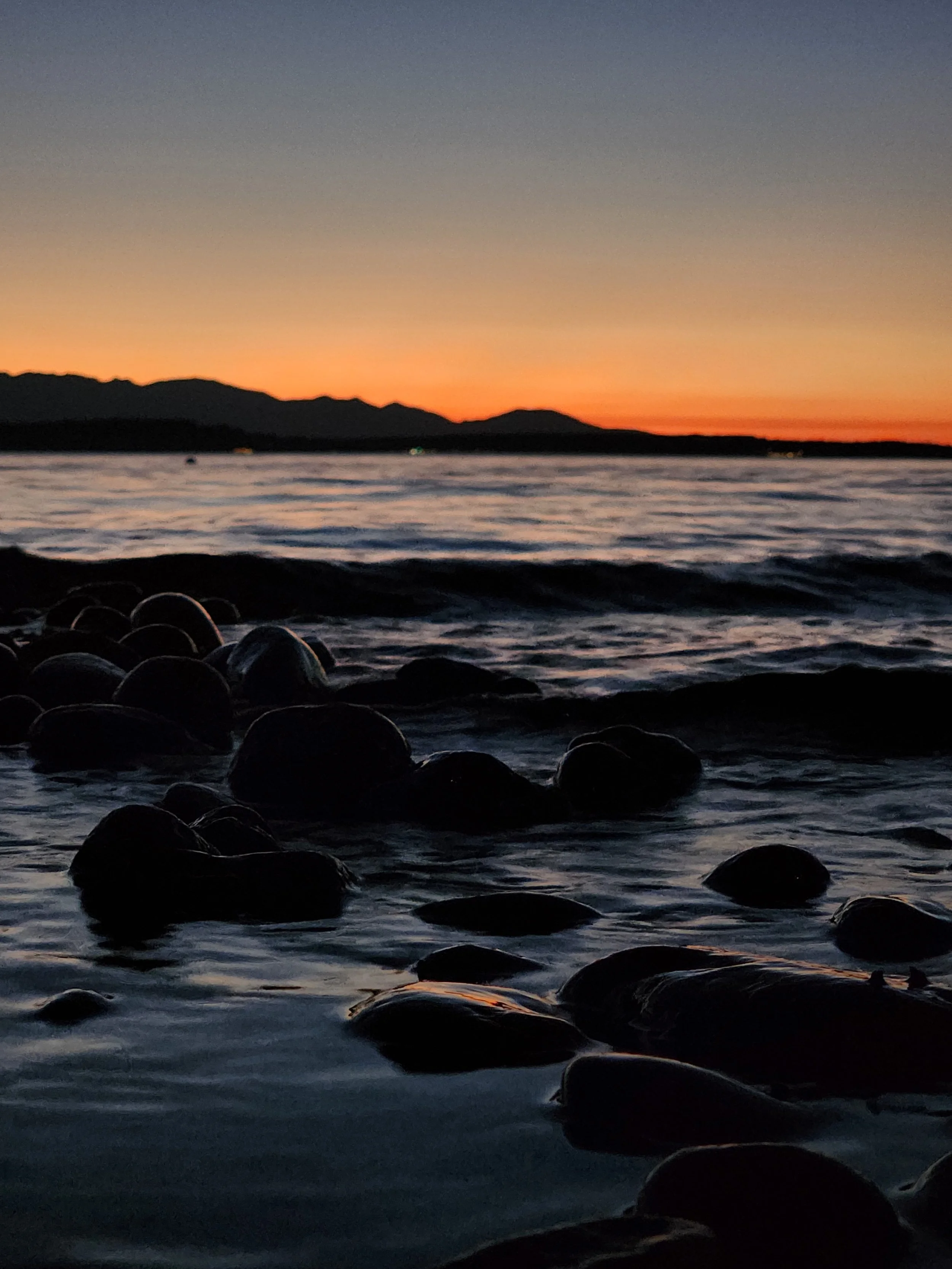 Sunset  over mountains in the background with water coming to shore over smooth rocks in the foreground