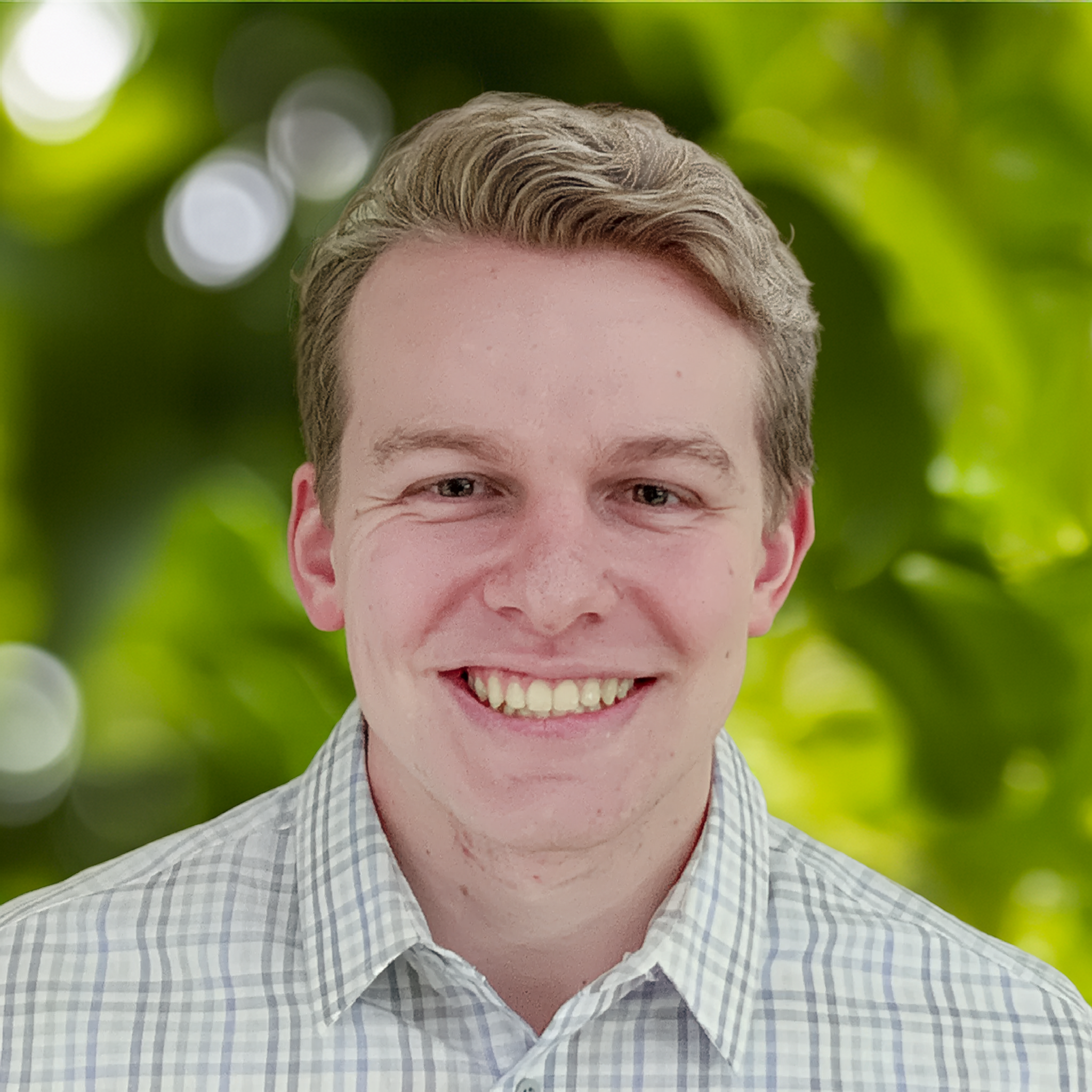 Headshot of a young man with light skin and red hair, smiling, wearing a checkered shirt, standing against a plain light-colored background.