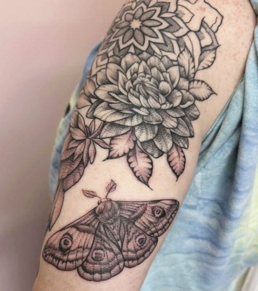 Black ink tattoo featuring a mandala pattern with skulls and floral design on skin, brick background.