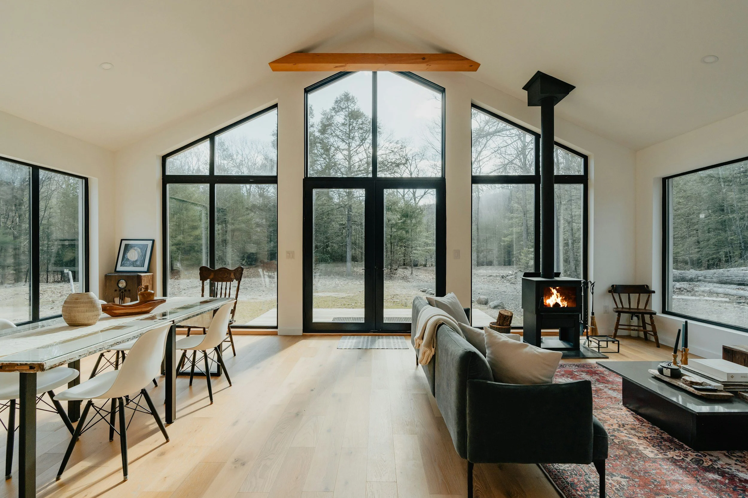 Modern living room with large floor-to-ceiling windows showing a wooded outdoor view, a black wood-burning stove with a visible fire, and minimalist furniture including a grey sectional sofa, a patterned area rug, and a wooden chair.