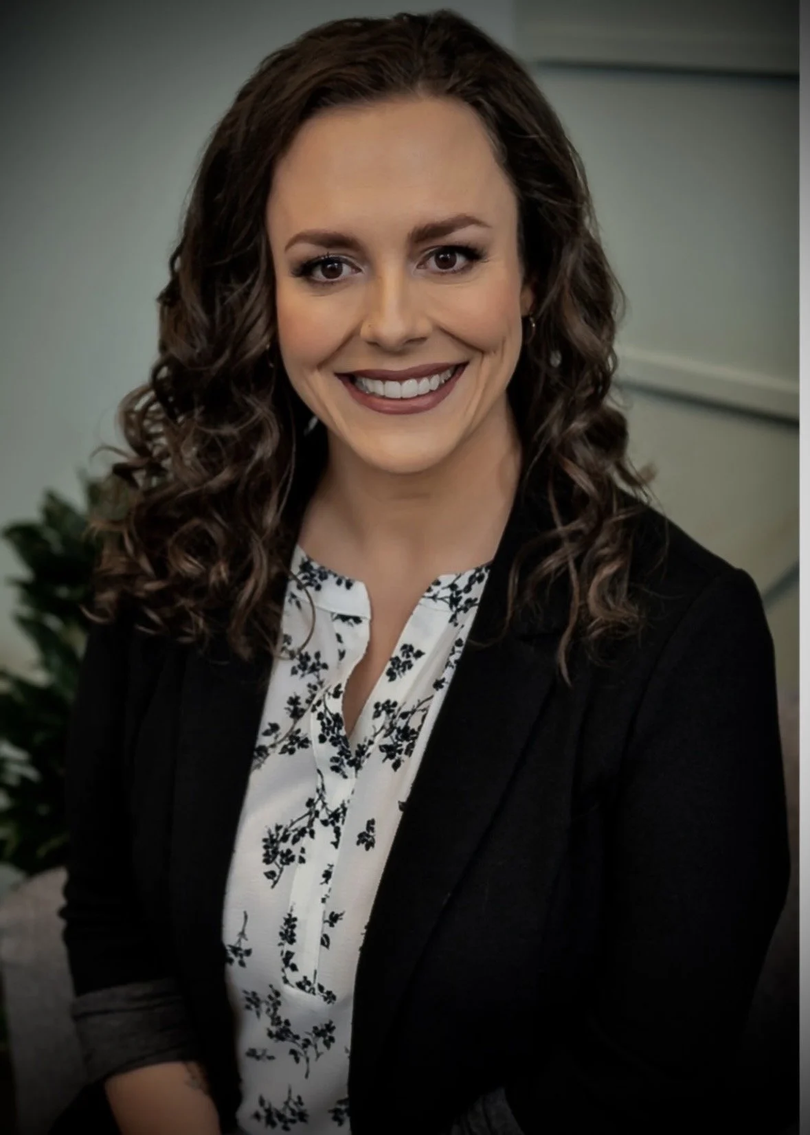 Deanna Woodroffe smiling in a professional headshot, wearing a black blazer over a white floral blouse, with long dark curly hair and a softly blurred office background.