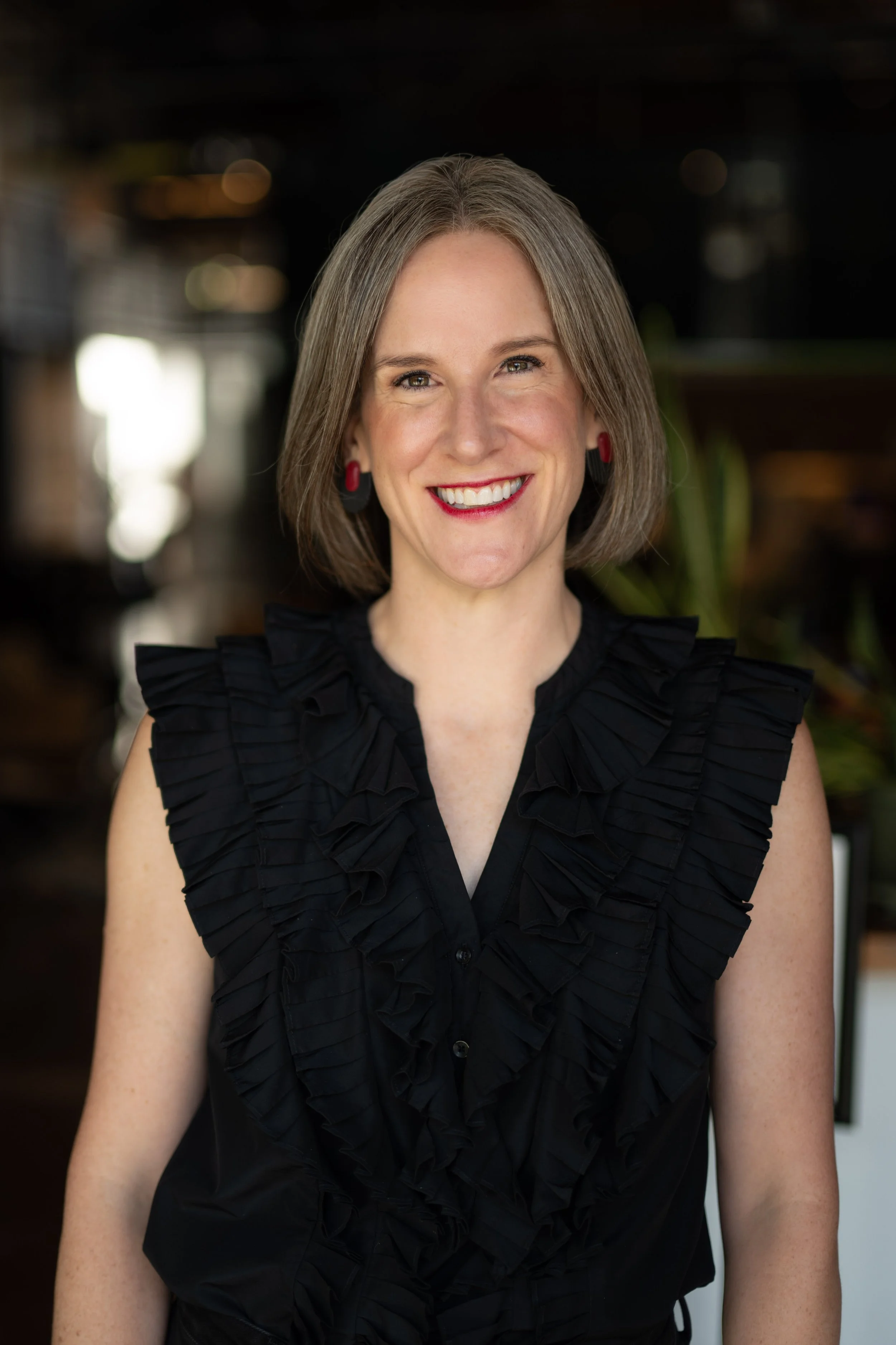 Elexis Schroder, founder and CEO of SchroderHaus, smiles for a professional portrait wearing a black ruffled blouse, with shoulder-length gray-brown hair and a softly blurred indoor background.