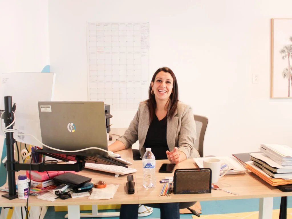 Ashley Ortiz, founder of Bella Vista Painting & Drywall, smiling at her desk in a bright office with a laptop, monitor, papers, and business supplies around her.