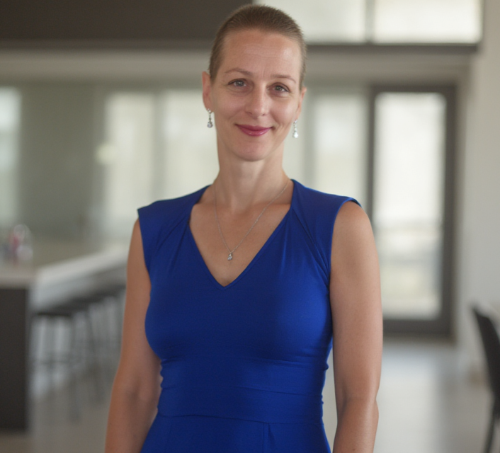 Natasha Golinsky, founder of On Purpose Projects, wearing a blue sleeveless dress and standing in a modern office setting with a blurred background.