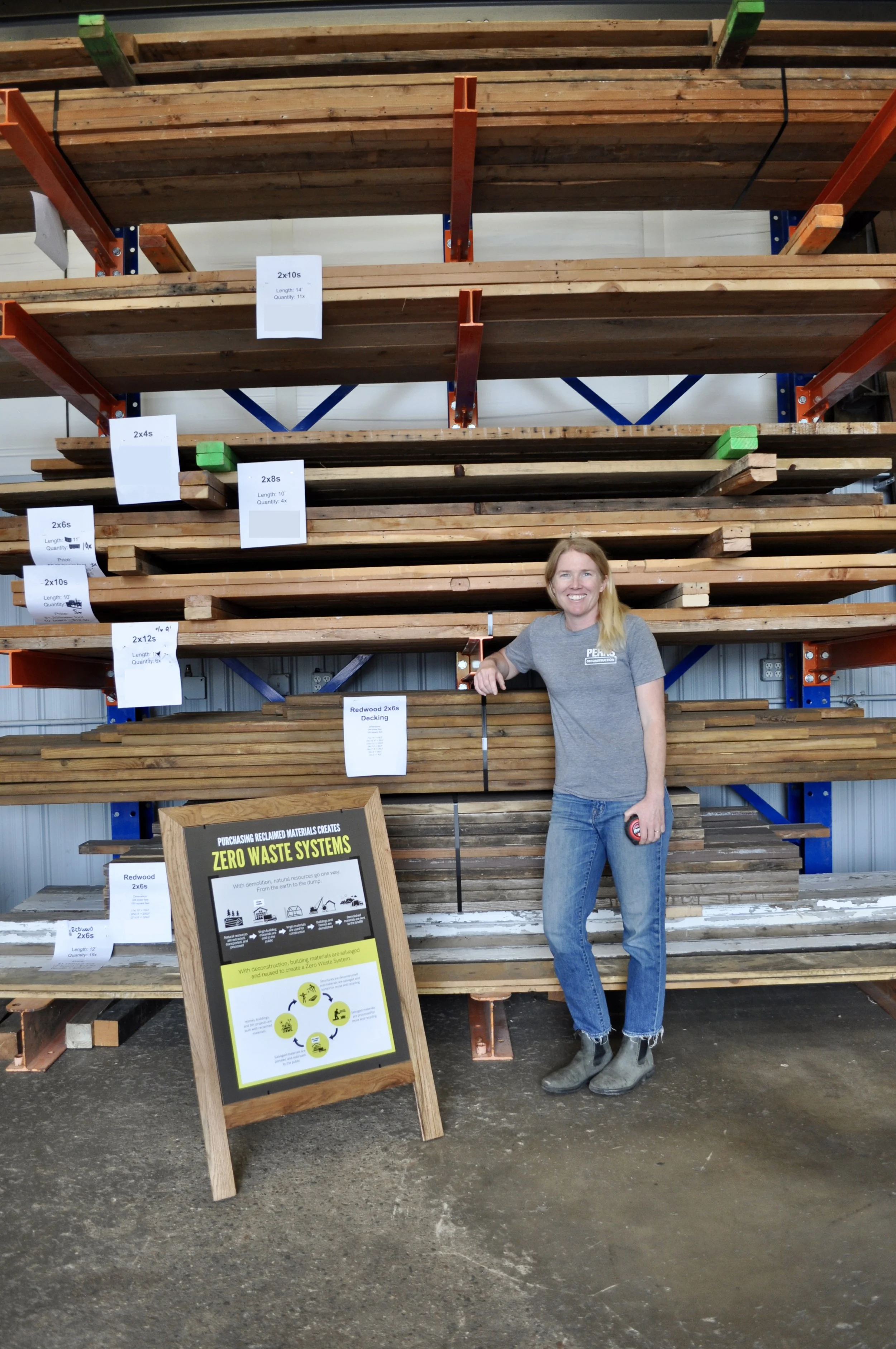 Anna Perks standing in a reclaimed lumber warehouse, smiling beside stacked shelves of salvaged wood beams and boards, with signage explaining zero waste systems and reclaimed building materials.
