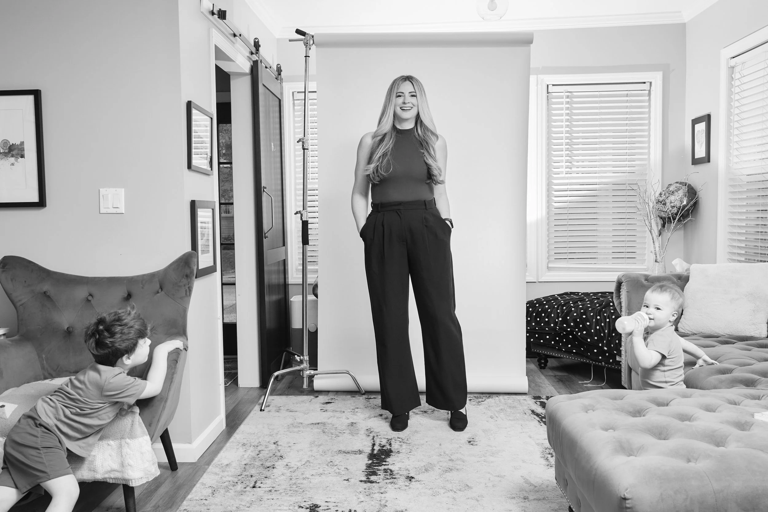 Black and white photo of Molly McCartan standing confidently in a home studio setup, smiling at the camera, with her two young children playing on either side of her.