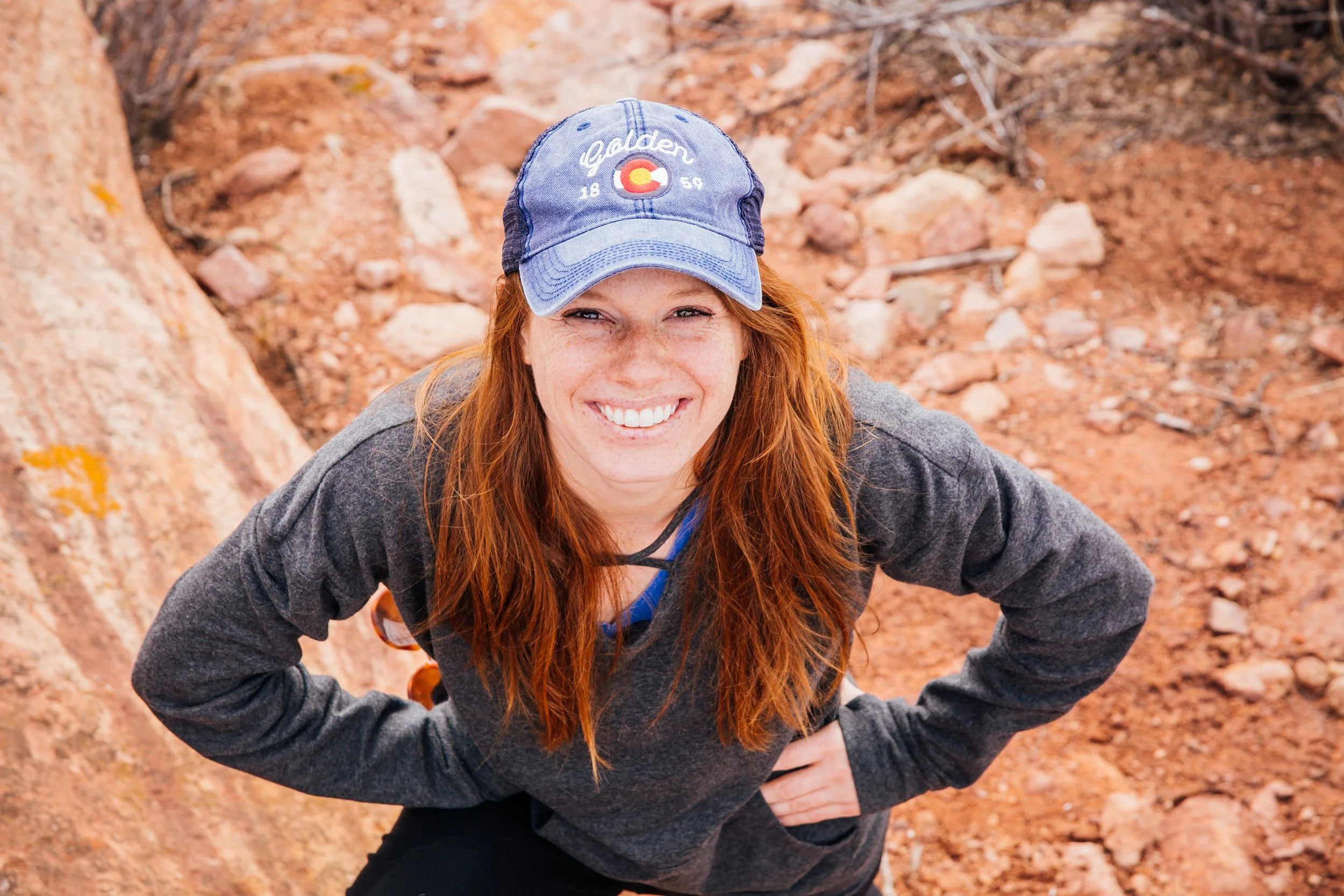 Rebecca Todd smiling outdoors on a rocky hiking trail in Colorado, wearing a blue Golden Colorado baseball cap and a dark long-sleeve top, looking up at the camera with hands on her hips.