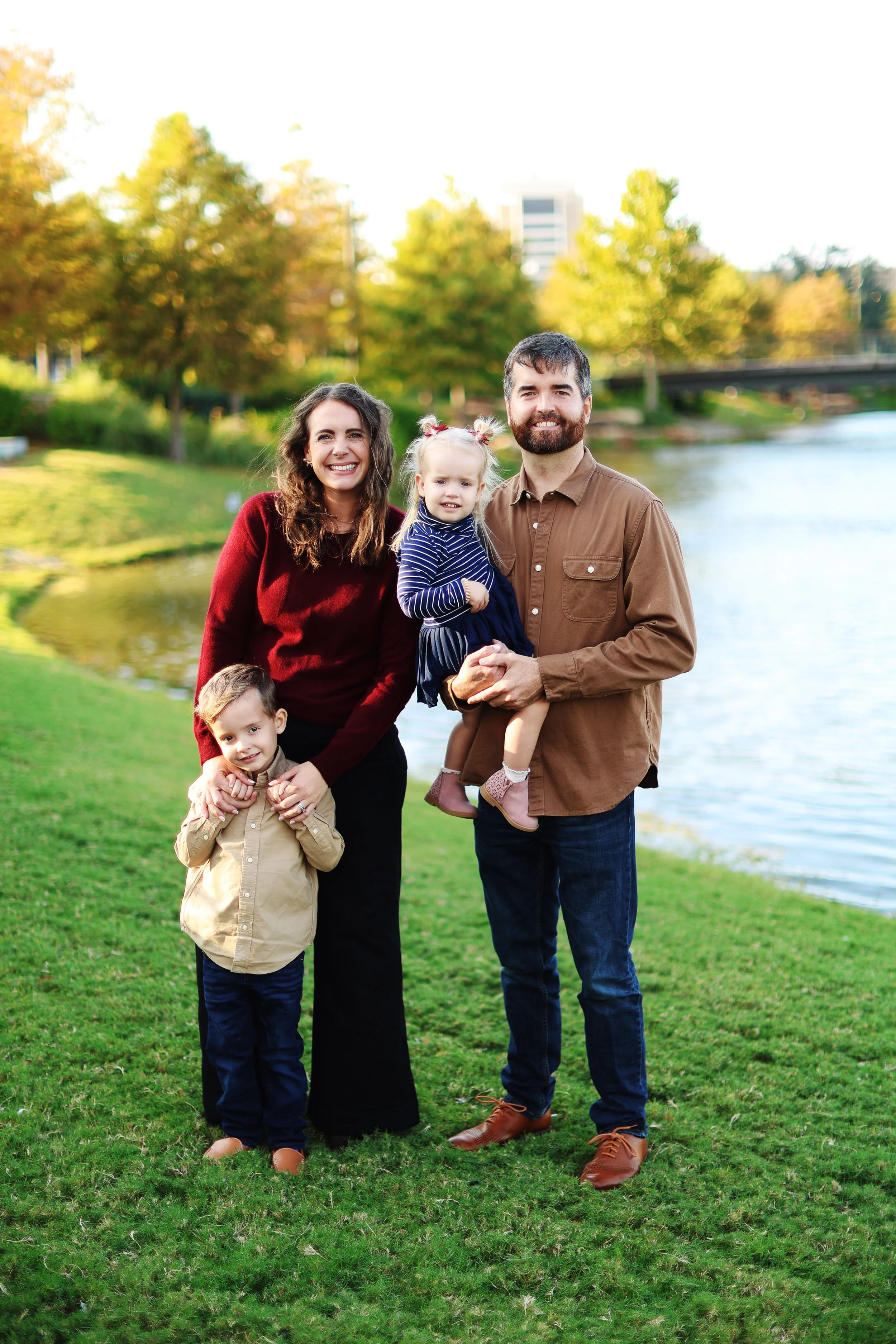Missy Wright standing outdoors in a park with her family of four by a river, smiling with her husband and two young children, with trees and a bridge in the background.