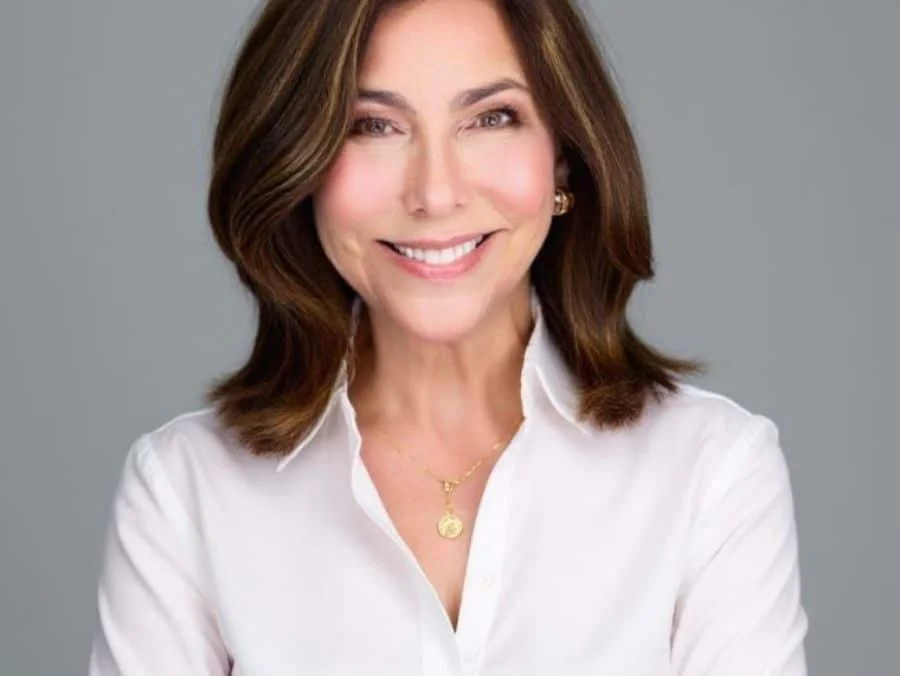 Professional headshot of Deborah Farone, smiling, wearing a white blouse and gold necklace against a neutral gray background.